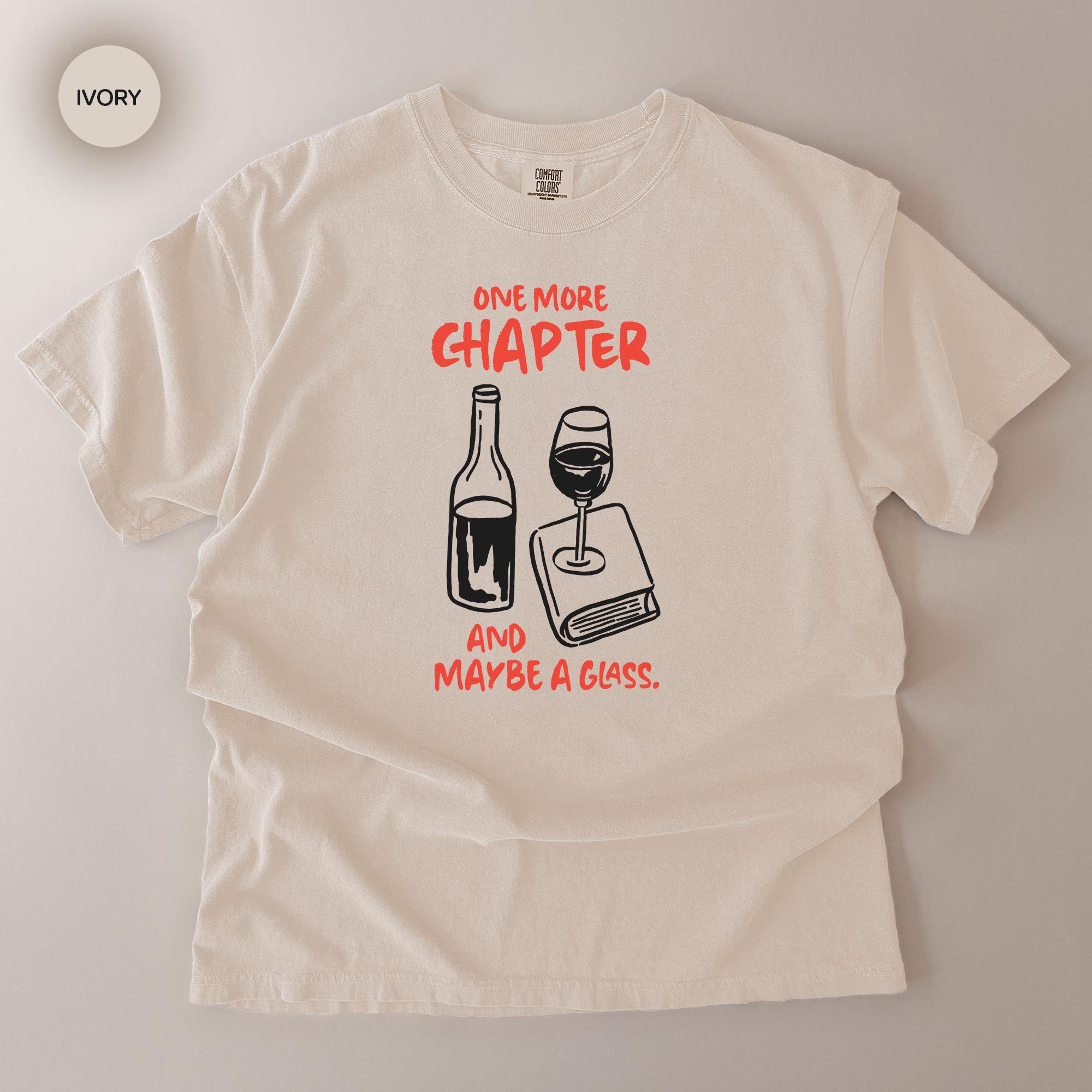 A white t-shirt with a graphic design featuring a wine bottle and a wine glass, along with the text "ONE MORE CHAPTER AND MAYBE A GLASS" printed on it.