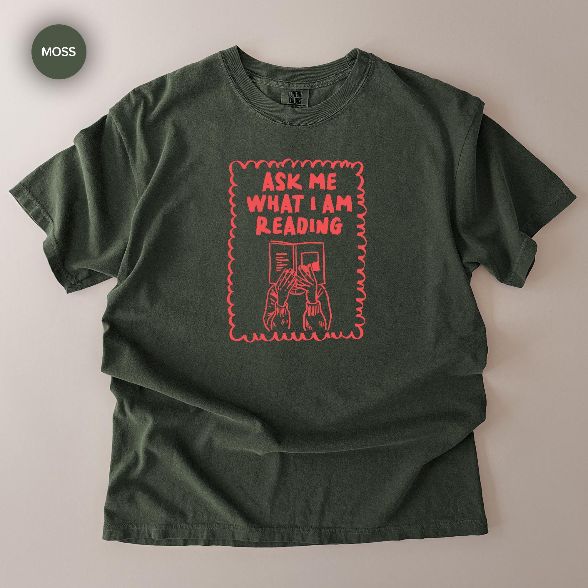 a green t-shirt with a graphic design on it. The design features a person reading a book, with the text "Ask me what I am reading" written above the person.
