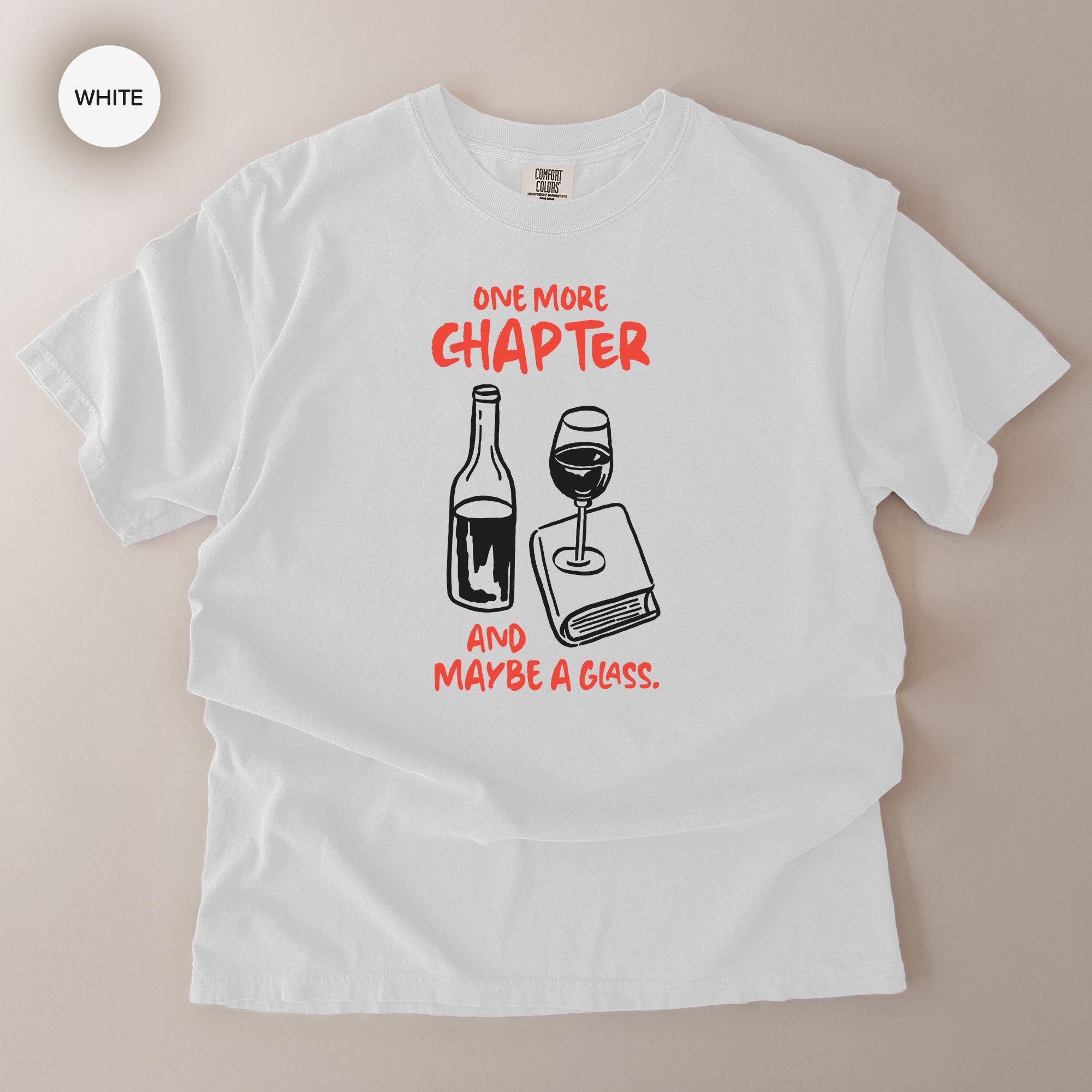 A white t-shirt with a graphic design depicting a wine bottle and a wine glass, along with the text "One more chapter and maybe a glass."