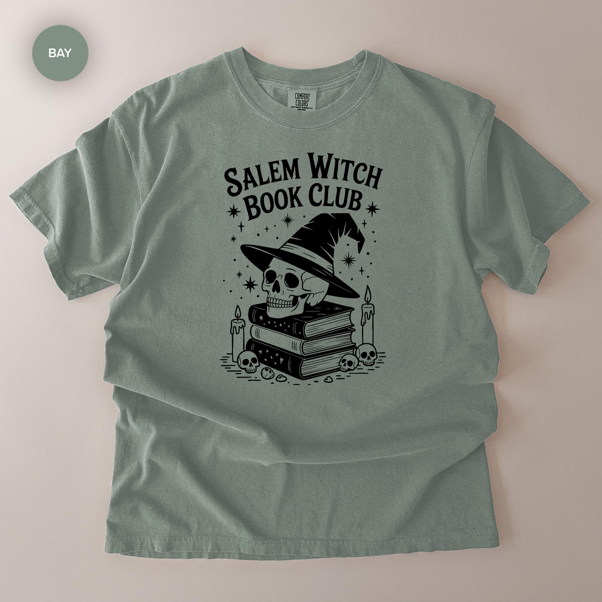 A gray t-shirt with a graphic design featuring a skull wearing a witch's hat, surrounded by books and candles, with the text "Salem Witch Book Club" printed on it.