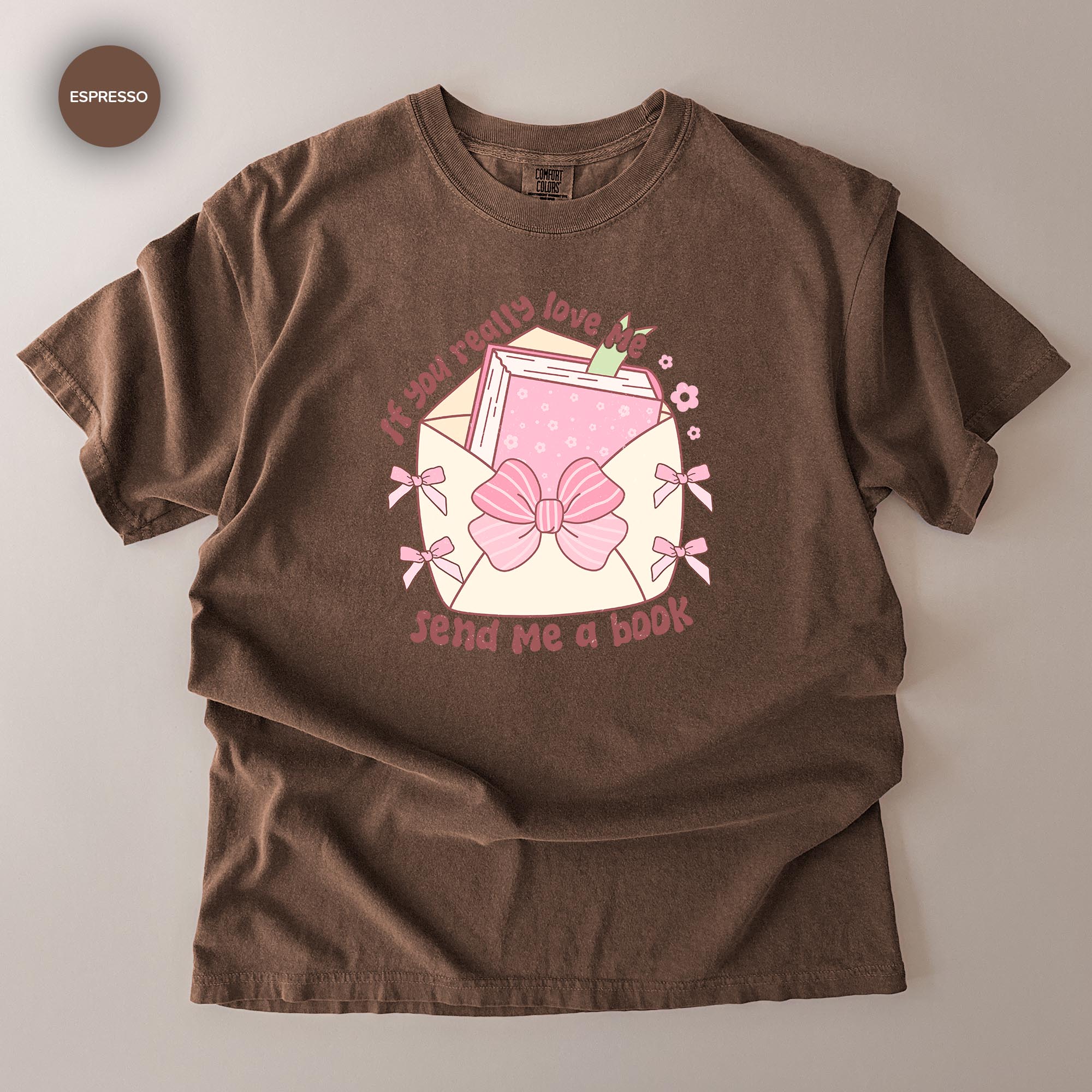 A brown t-shirt with a graphic design featuring a pink bow, a book, and the text "If you really love send me a book".