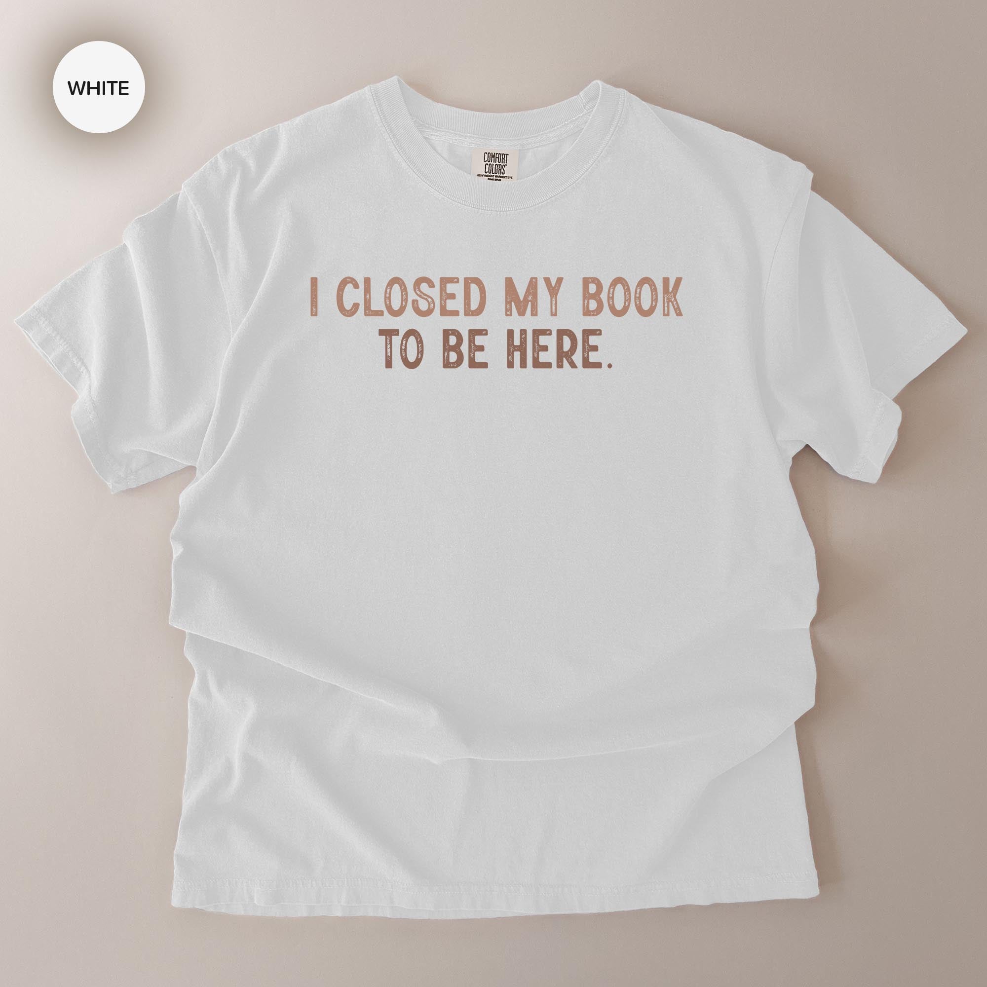 A white t-shirt with the text "I CLOSED MY BOOK TO BE HERE" printed on it.
