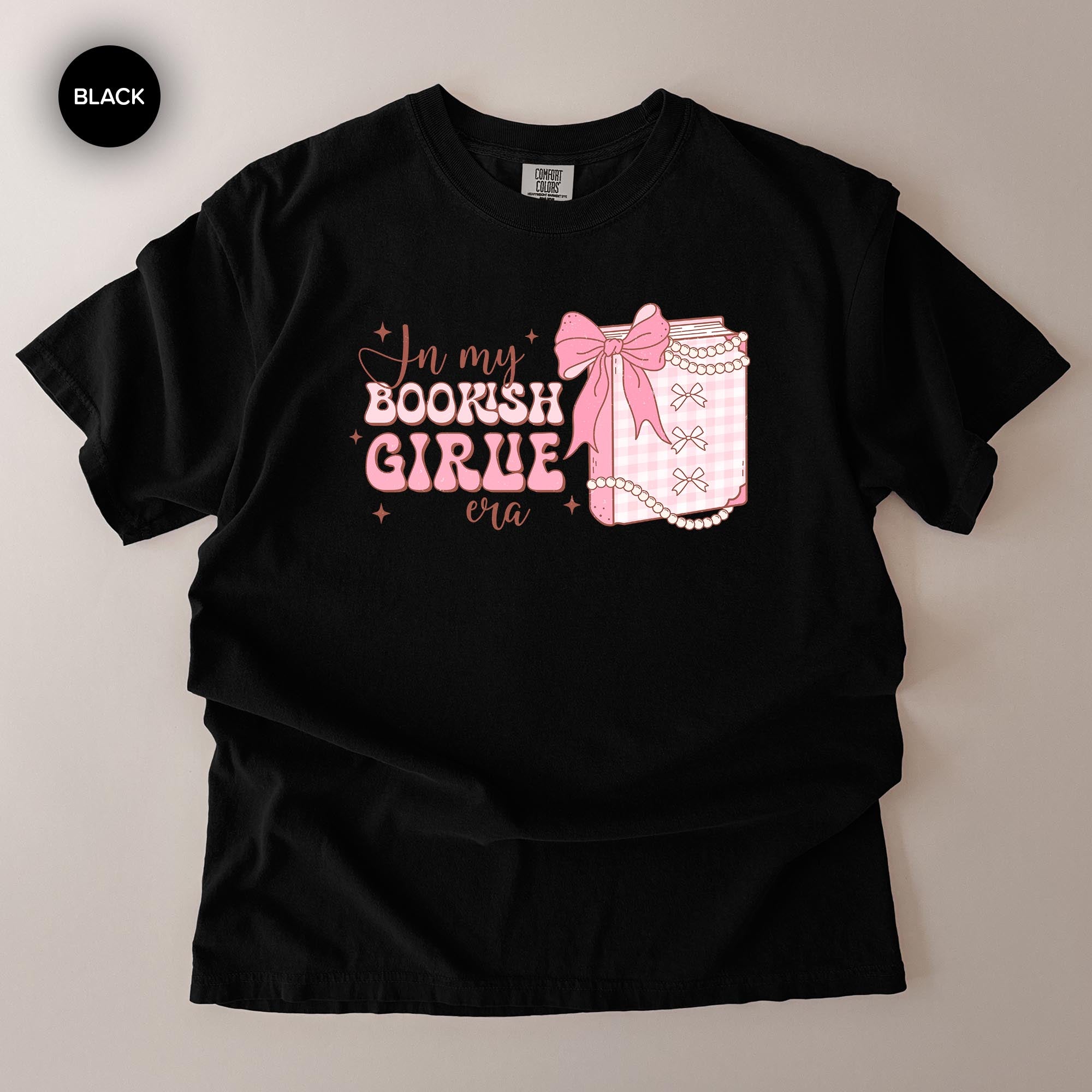 A black t-shirt with a graphic design featuring a bow, pearls, and text that reads "In my BOOKISH GIRLE era".
