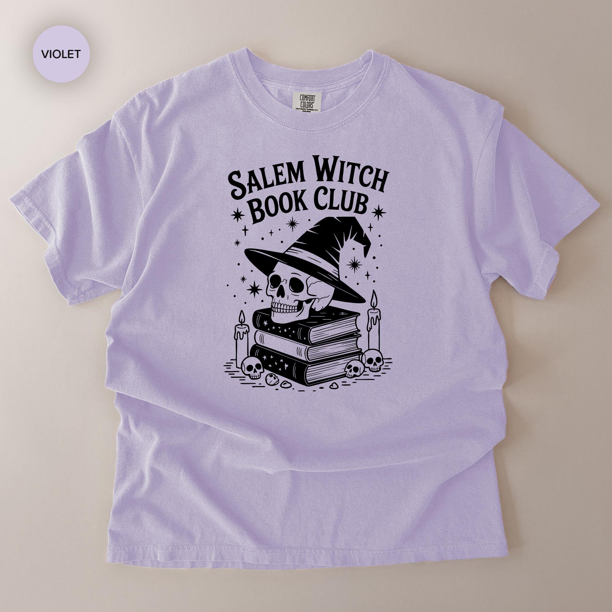 A purple t-shirt with a graphic design featuring a skull wearing a witch's hat, surrounded by books and candles, with the text "Salem Witch Book Club" printed on it.