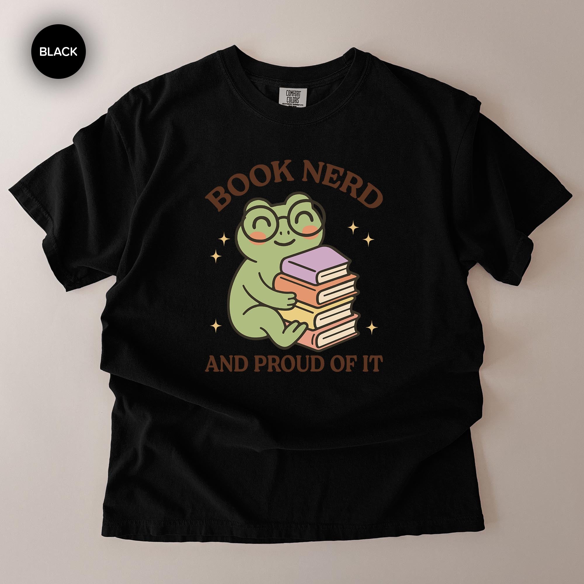 A black t-shirt with a graphic design featuring a smiling frog holding a stack of books, with the text "BOOK NERD AND PROUD OF IT" above the frog.