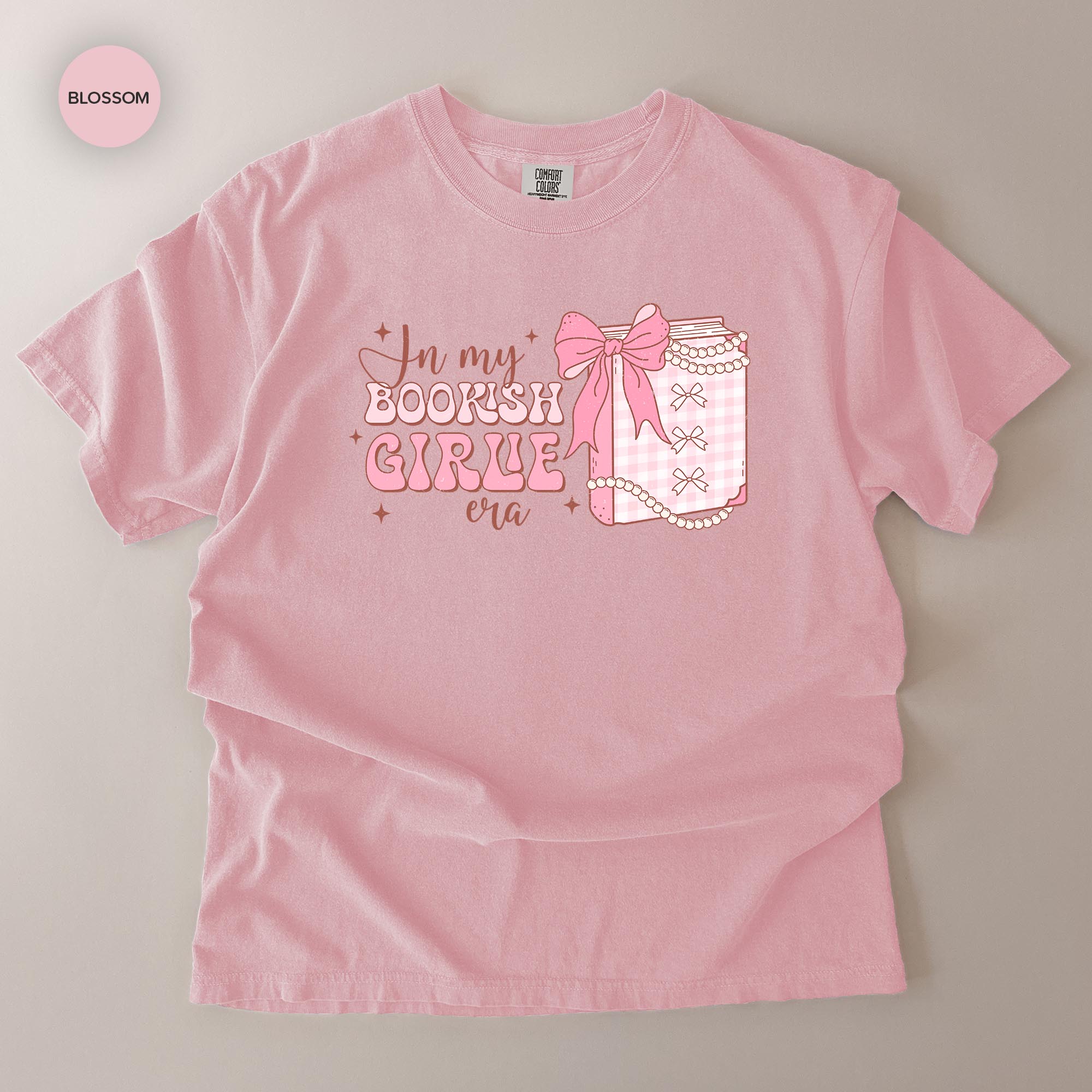 A pink t-shirt with a graphic design that says "In my BOOKISH GIRLE era" and features a bow and a small book.