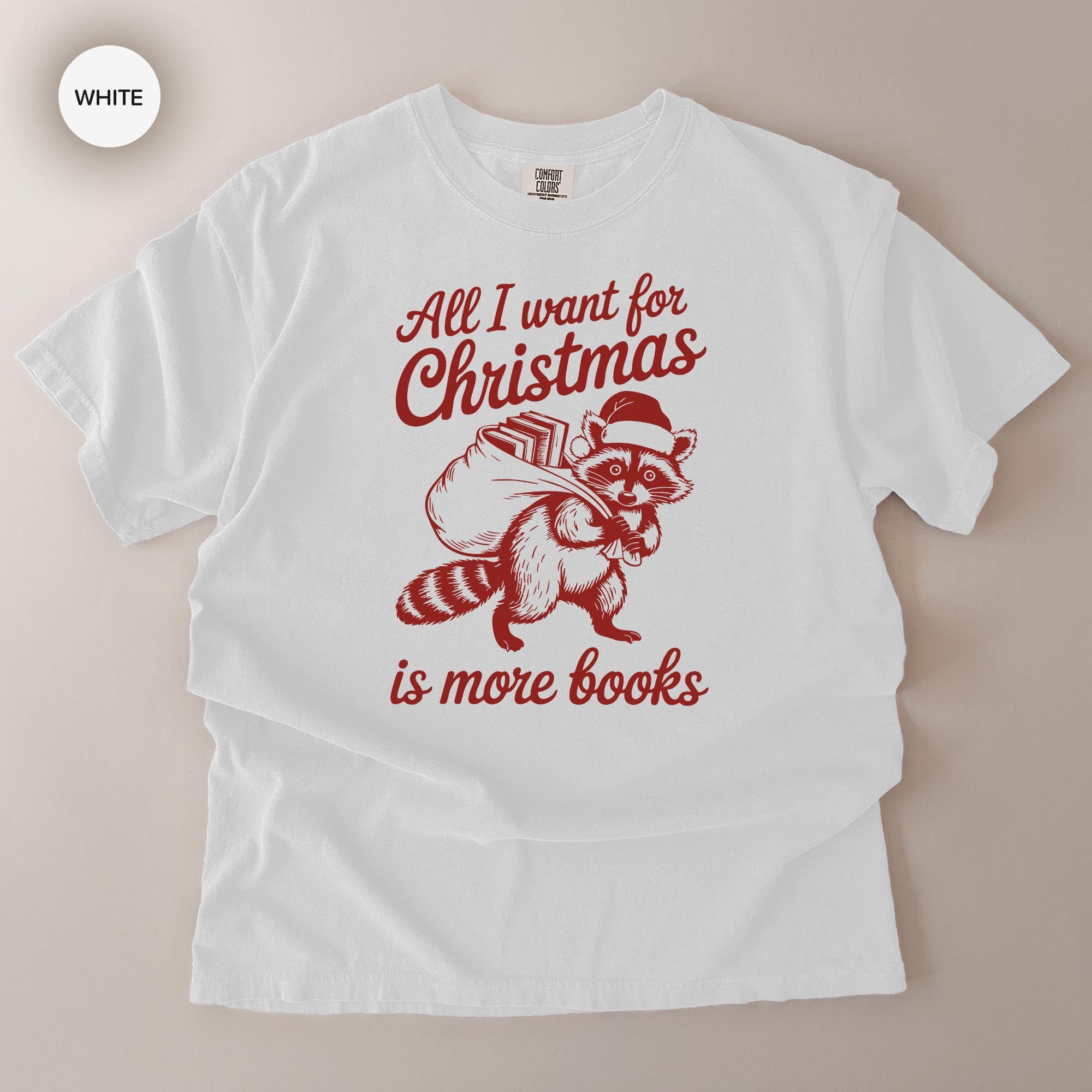 A white t-shirt with a red graphic design depicting a raccoon carrying a sack of books and the text "All I want for Christmas is more books".