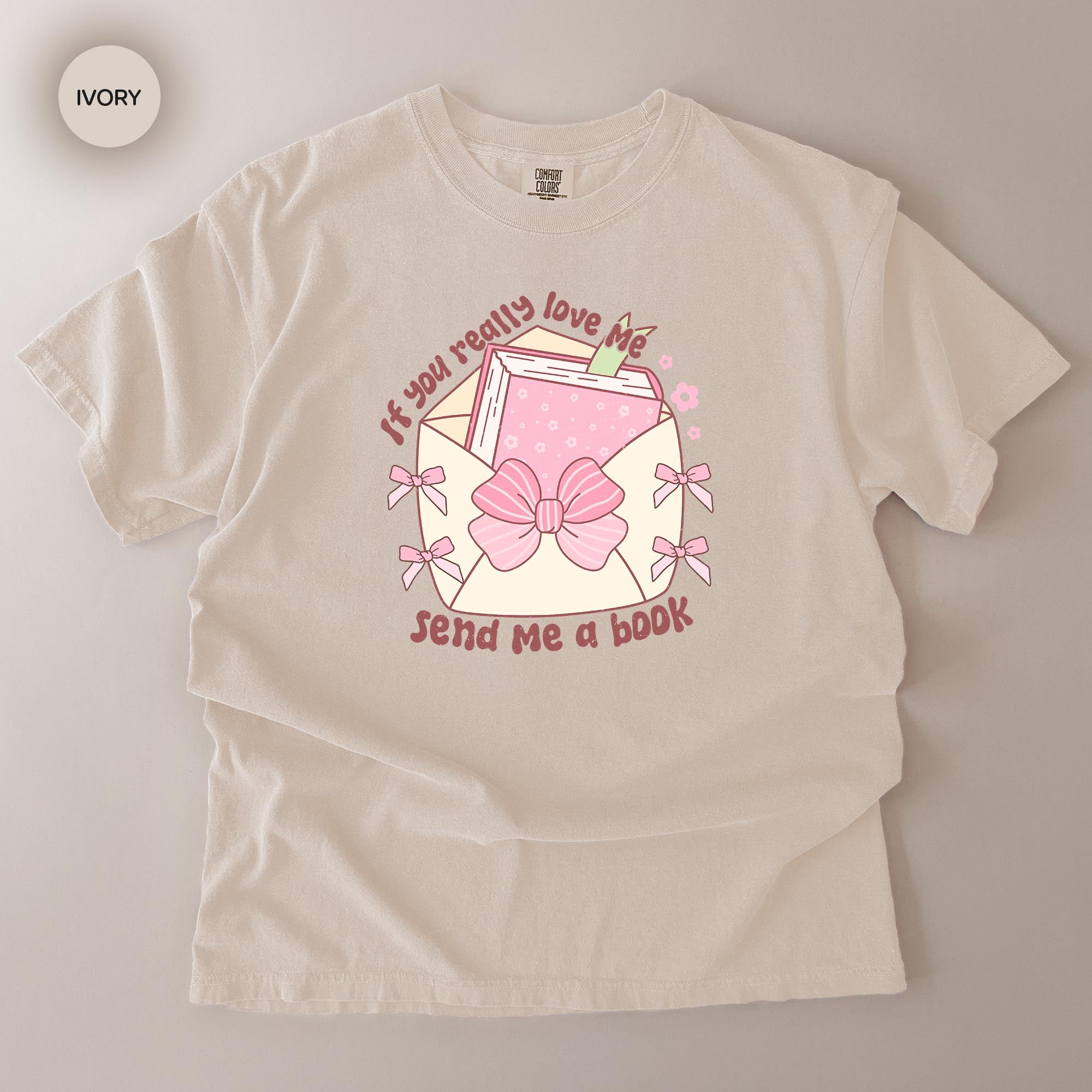 A white t-shirt with a graphic design featuring a pink bow, a book, and the text "If you really love me, send me a book".