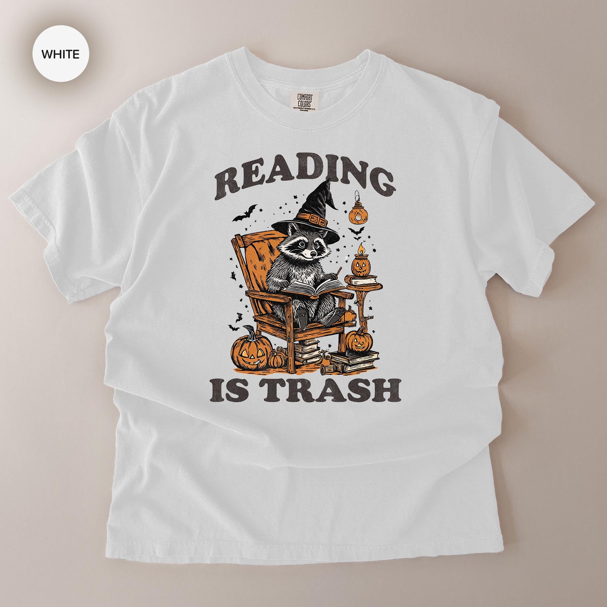 A white t-shirt with a graphic design featuring a raccoon wearing a witch's hat and holding a book, surrounded by Halloween-themed elements such as pumpkins and bats.
