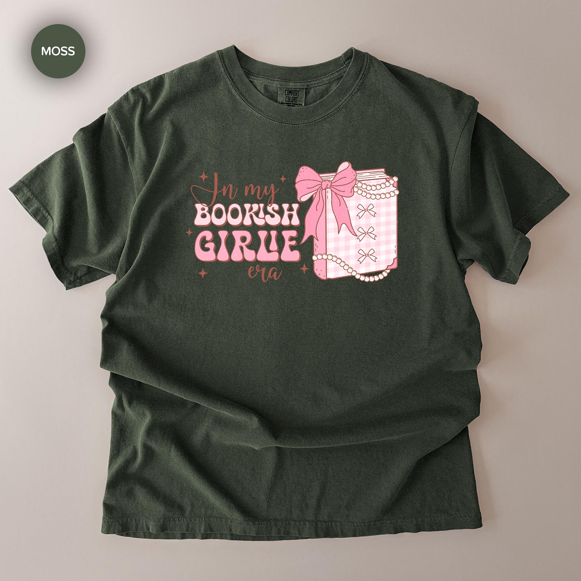 A dark green t-shirt with a graphic design featuring a bow, pearls, and text that reads "In my BOOKISH GIRLE era".