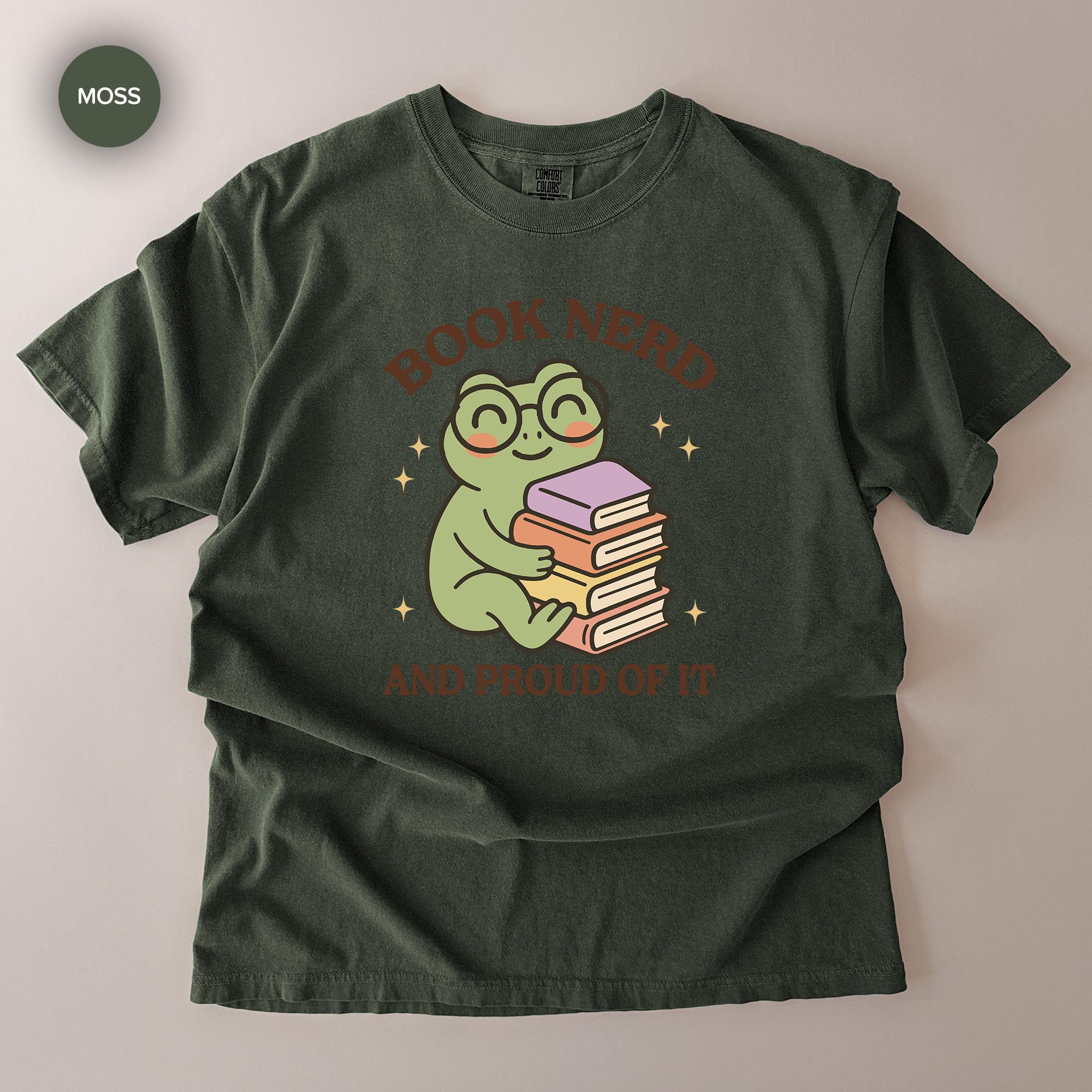 A green t-shirt with a graphic design featuring a frog holding a stack of books, surrounded by stars. The text above the frog reads "BOOK NERD" and below the frog reads "AND PROUD OF IT".