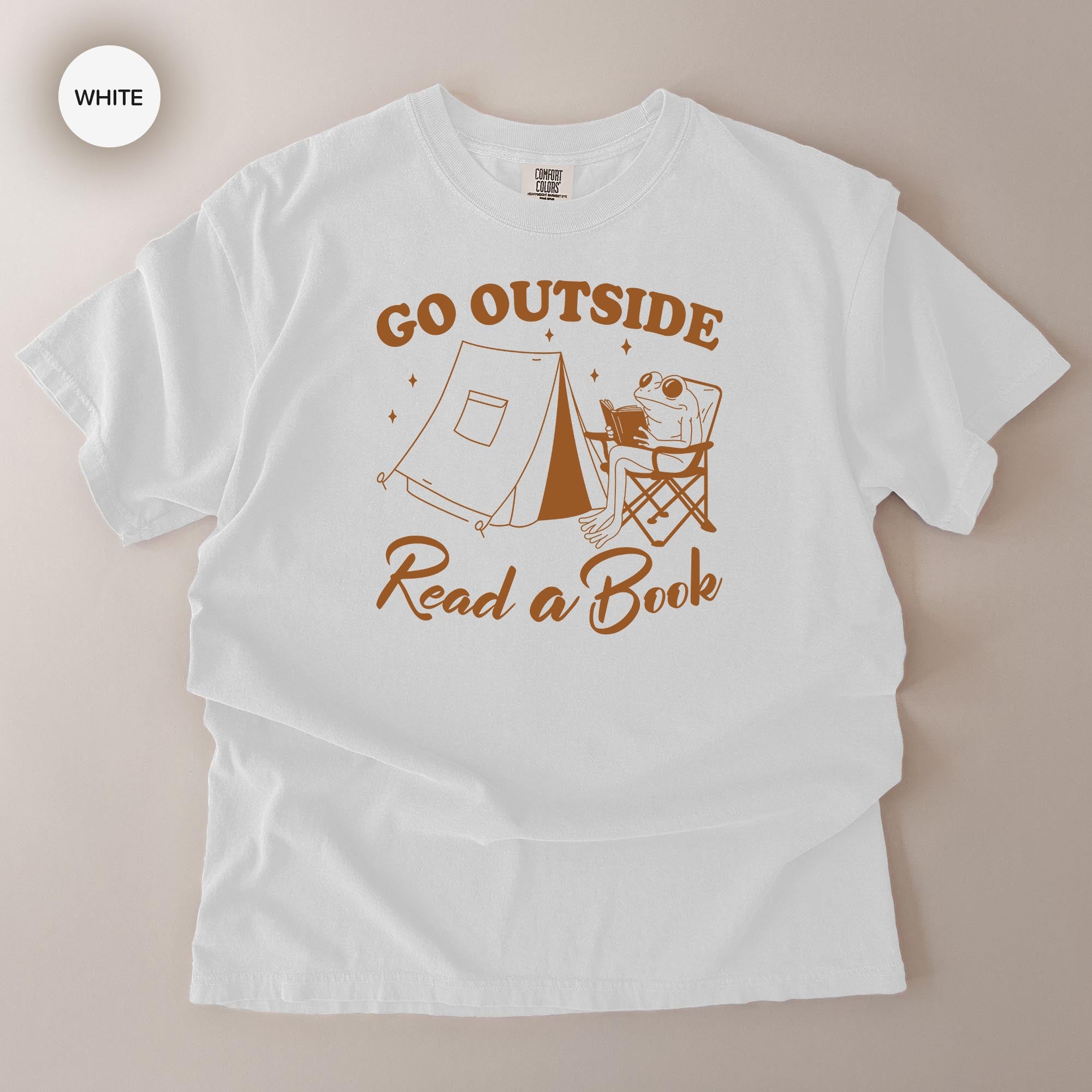 A white t-shirt with a graphic design that says "Go Outside Read a Book" and features a frog sitting in a chair next to a tent.