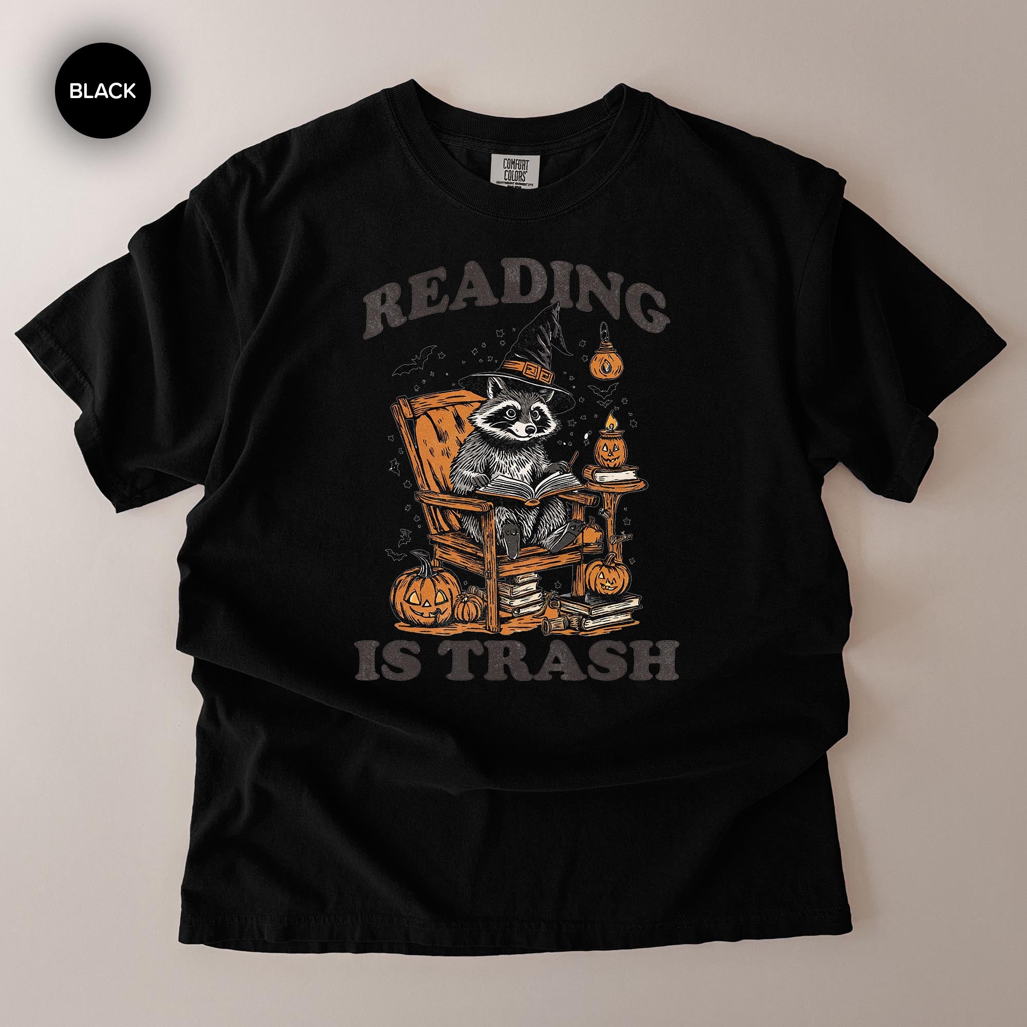 A black t-shirt with a graphic design depicting a raccoon sitting in a chair and reading a book, surrounded by Halloween-themed elements such as pumpkins and bats.