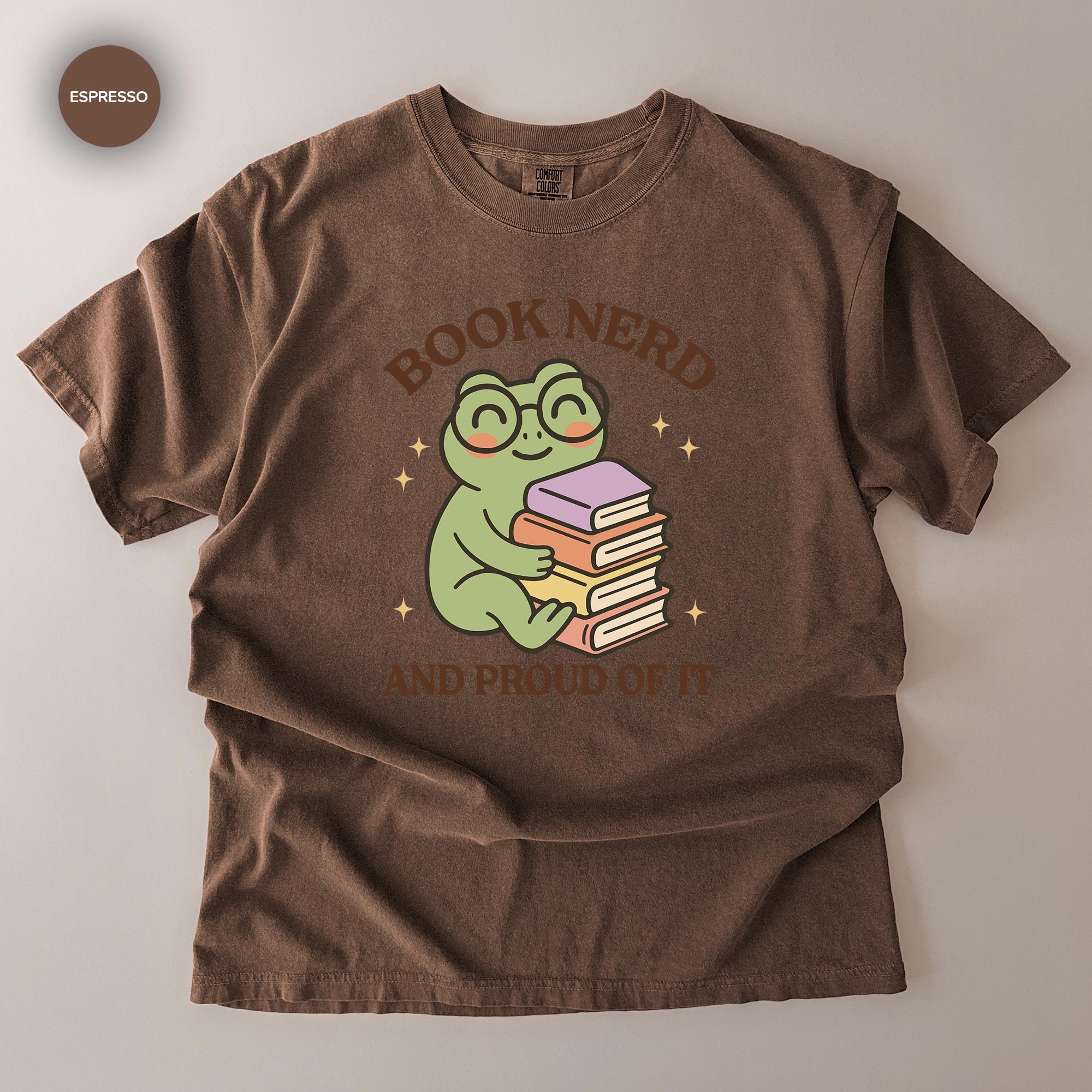 A brown t-shirt with a graphic design featuring a smiling frog holding a stack of books, along with the text "BOOK NERD AND PROUD OF IT".