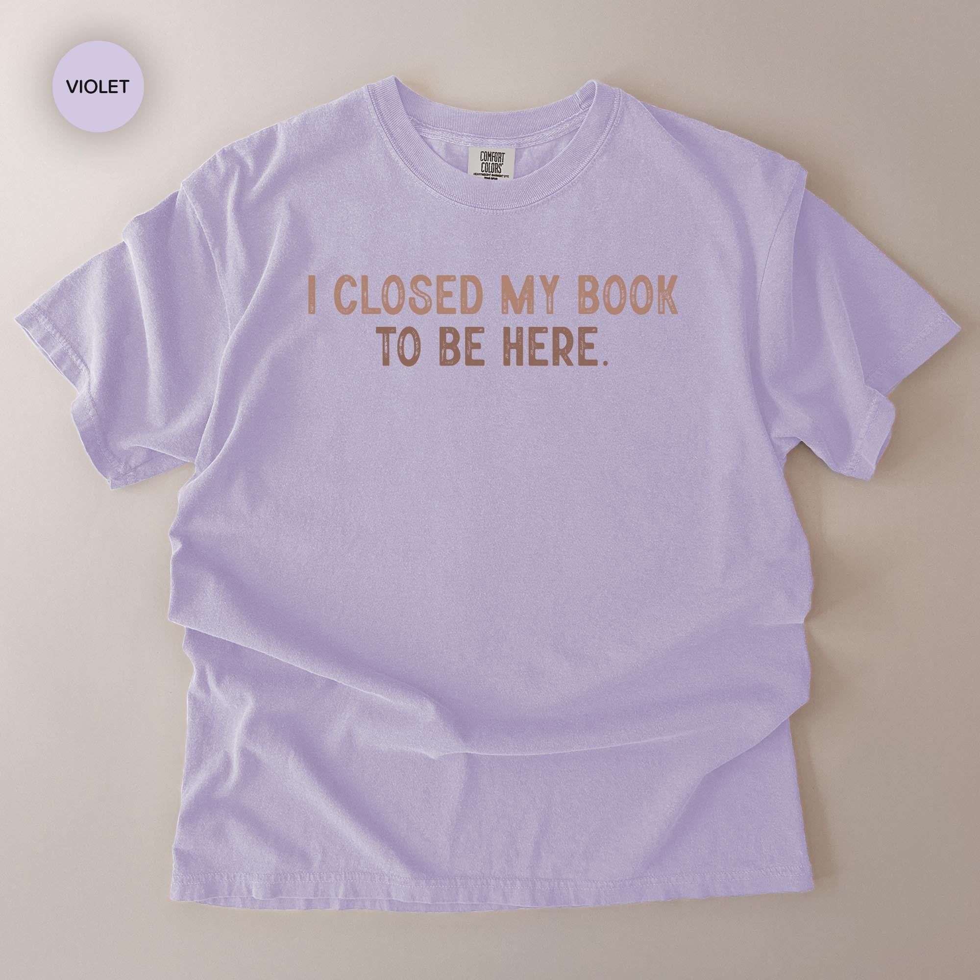 A light purple t-shirt with the text "I CLOSED MY BOOK TO BE HERE" printed on it.