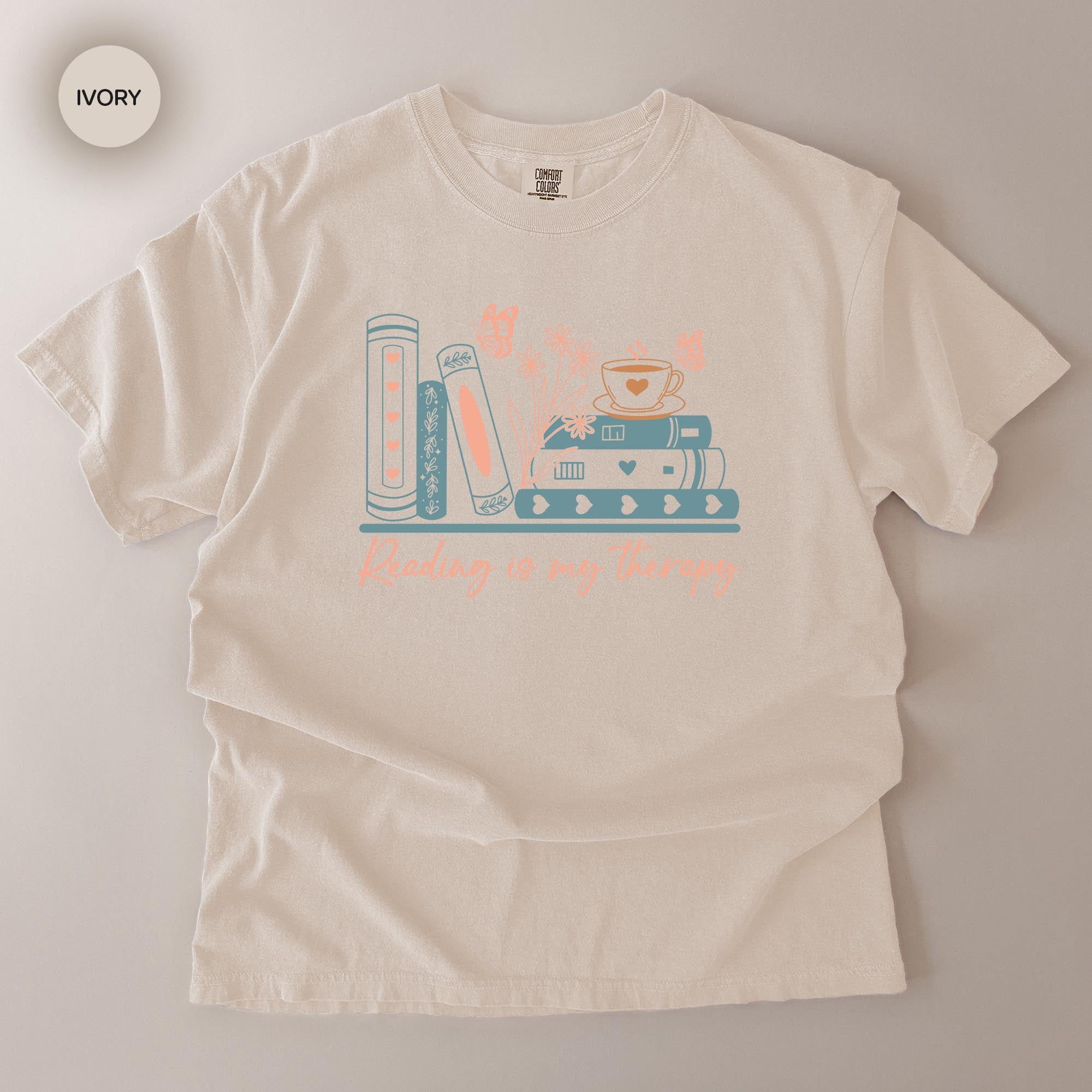 A white t-shirt with a graphic design featuring a stack of books and a cup of tea, along with the text "Reading is my therapy" printed on it.
