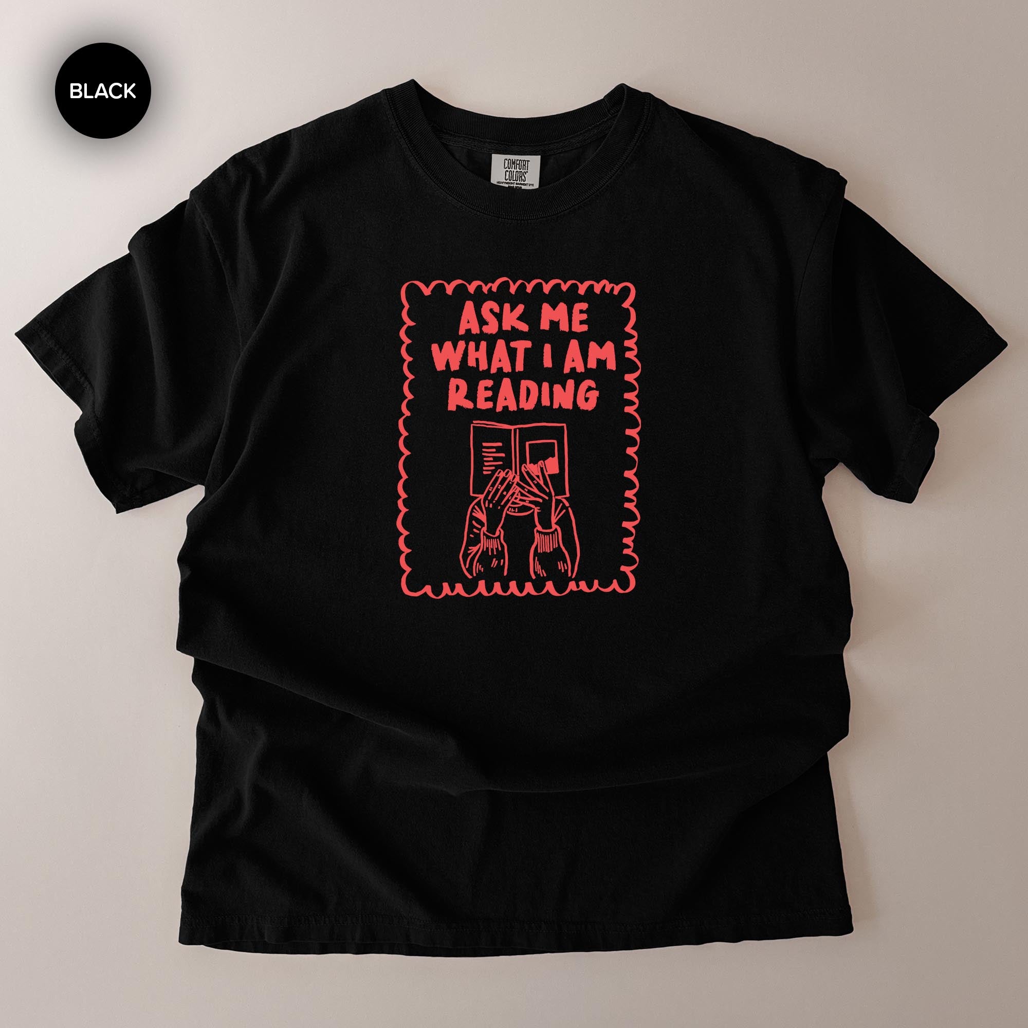 The image is a black t-shirt with a graphic design on it. The design features a person reading a book, with the text "Ask me what I am reading" written above the person.