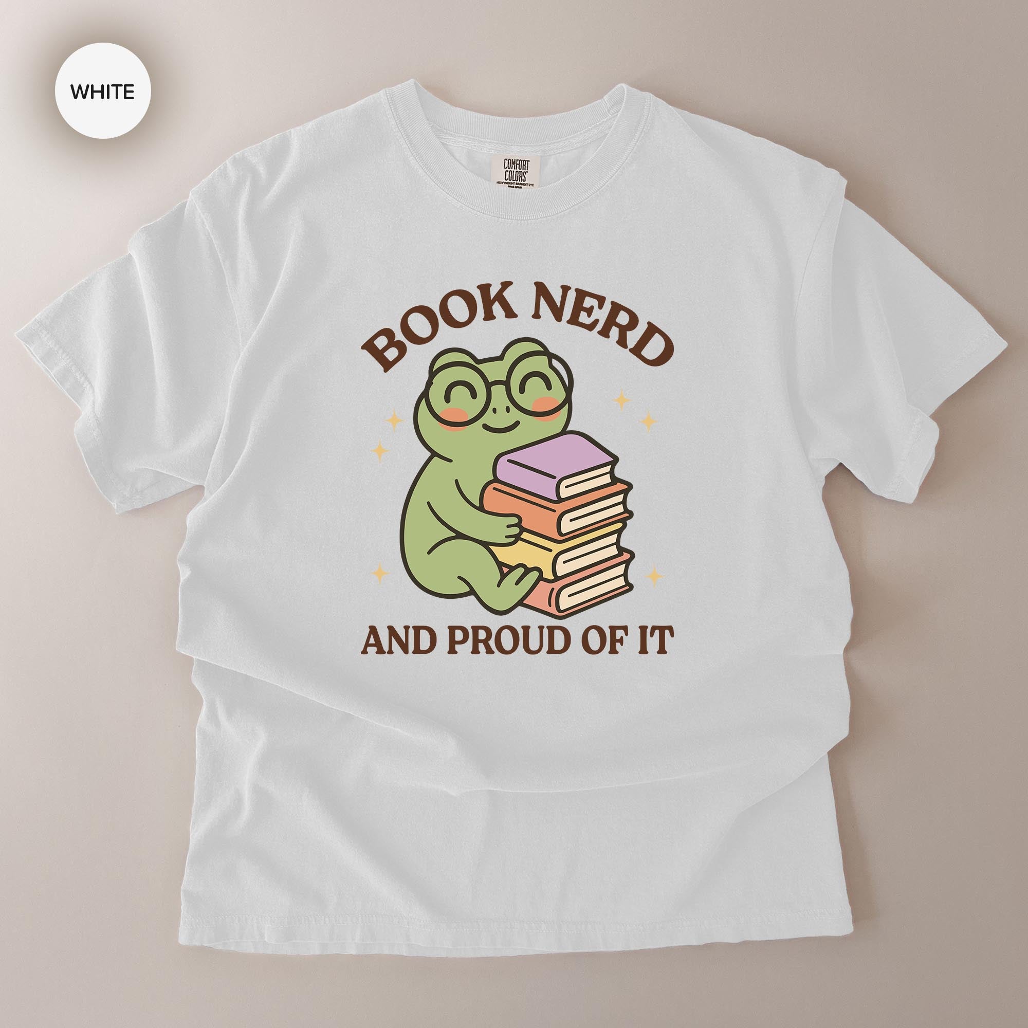 A white t-shirt with a graphic design featuring a frog wearing glasses and holding a stack of books, along with the text "BOOK NERD AND PROUD OF IT".