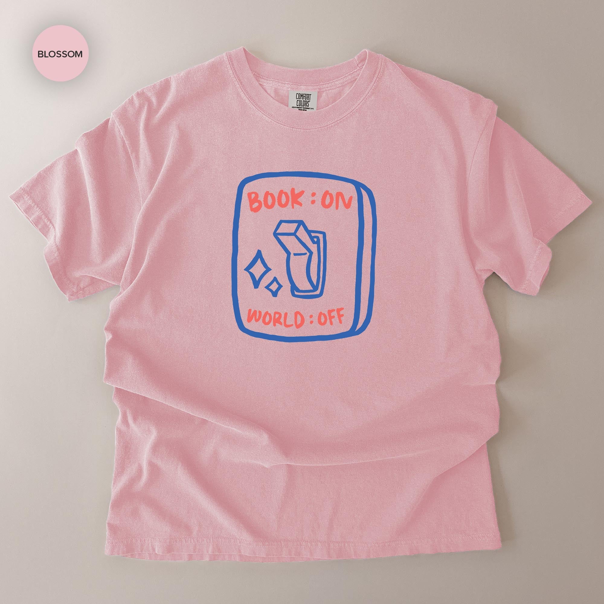 A pink t-shirt with a graphic design on it, featuring a blue rectangle with a white book and the text "BOOK ON WORLD+OFF" printed on it.