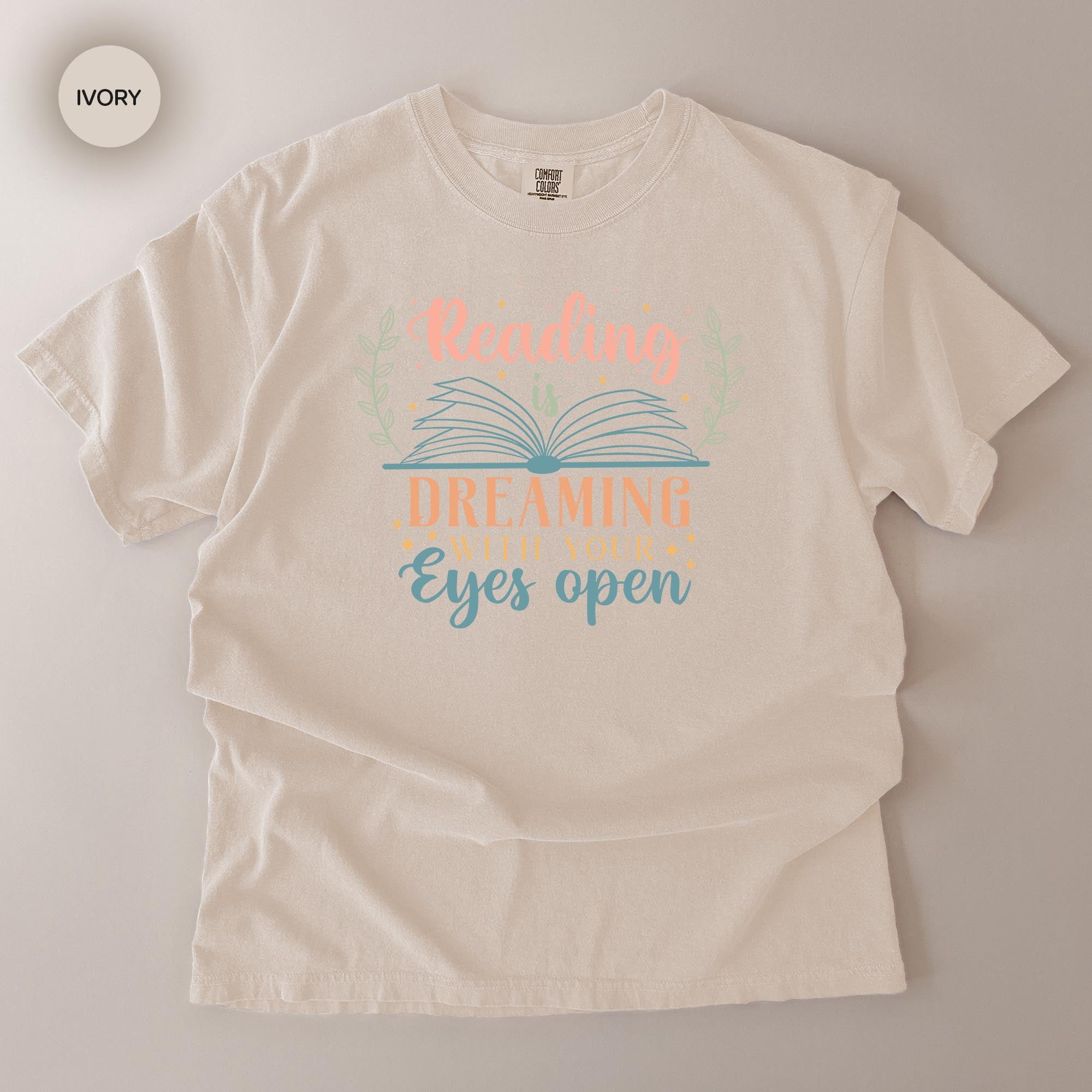 A white t-shirt with a printed design that says "Reading Dreaming With Your Eyes Open" in a colorful, stylized font.