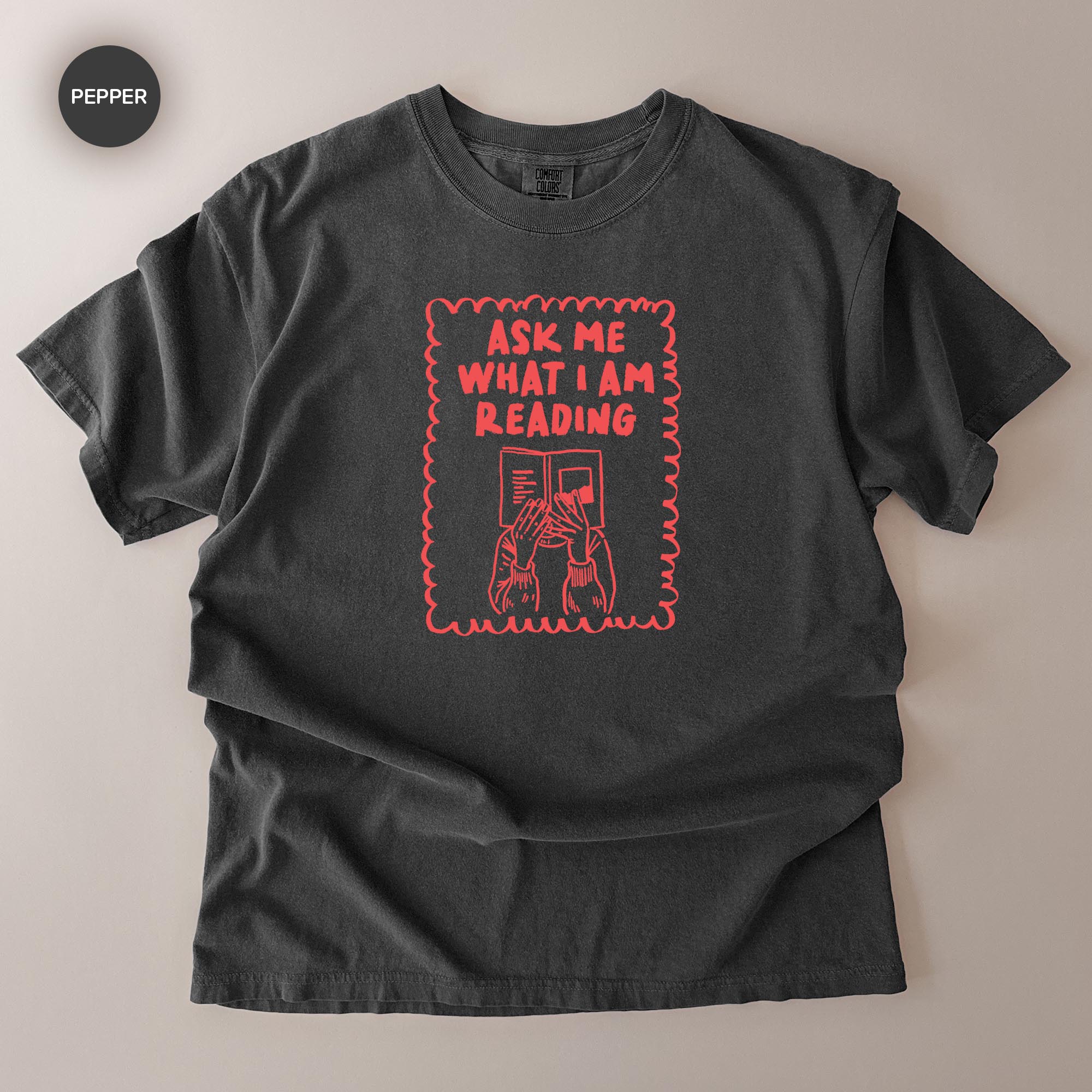 a black t-shirt with a red graphic design on it. The design includes a person reading a book and the text "Ask me what I am reading".