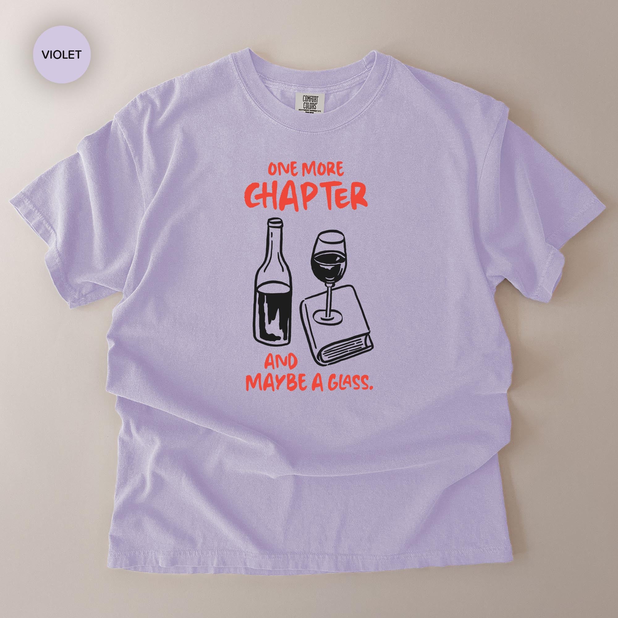 A purple t-shirt with a graphic design depicting a wine glass and a book, accompanied by the text "One more chapter and maybe a glass."