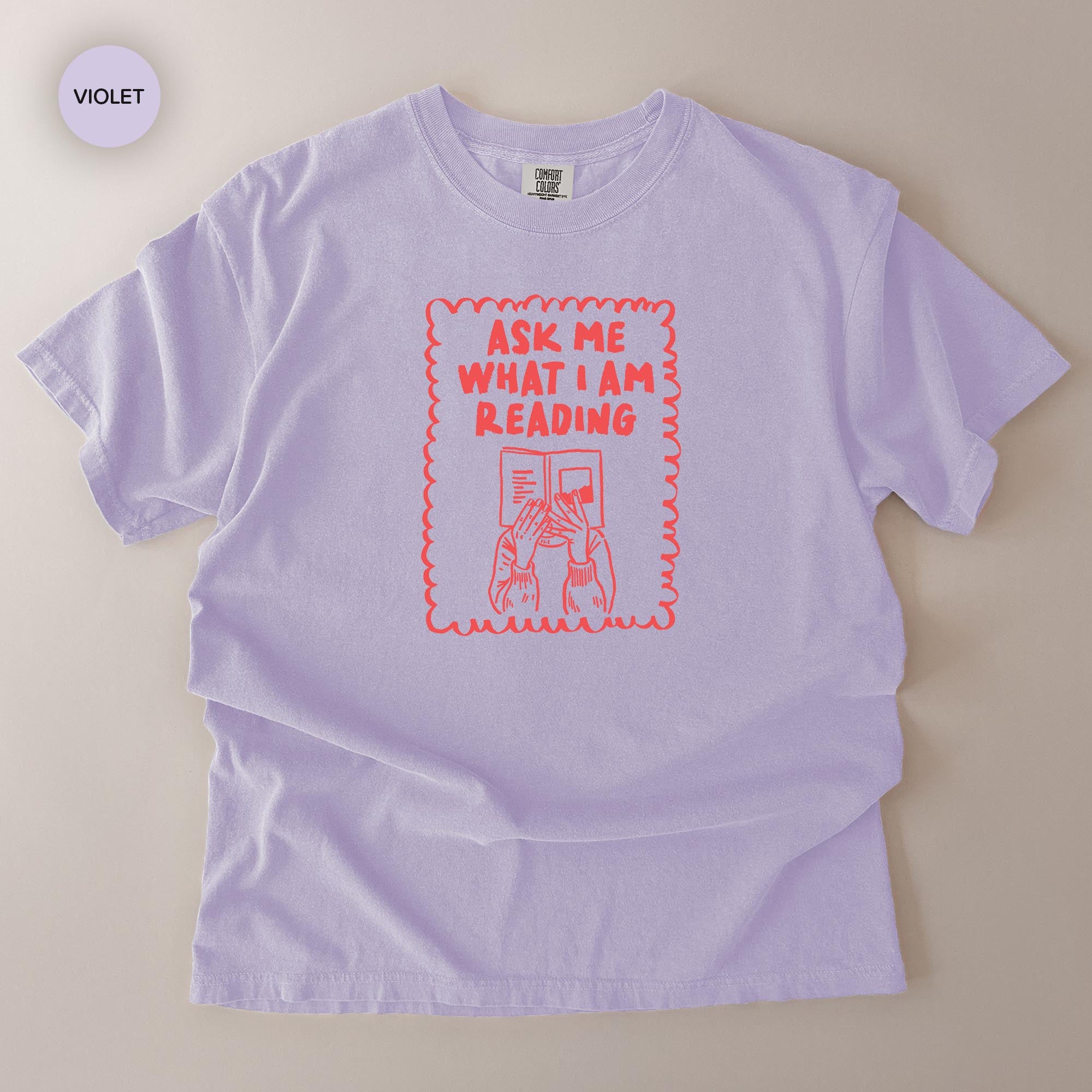 a purple t-shirt with a graphic design on it. The design includes a person reading a book and the text "Ask me what I am reading".