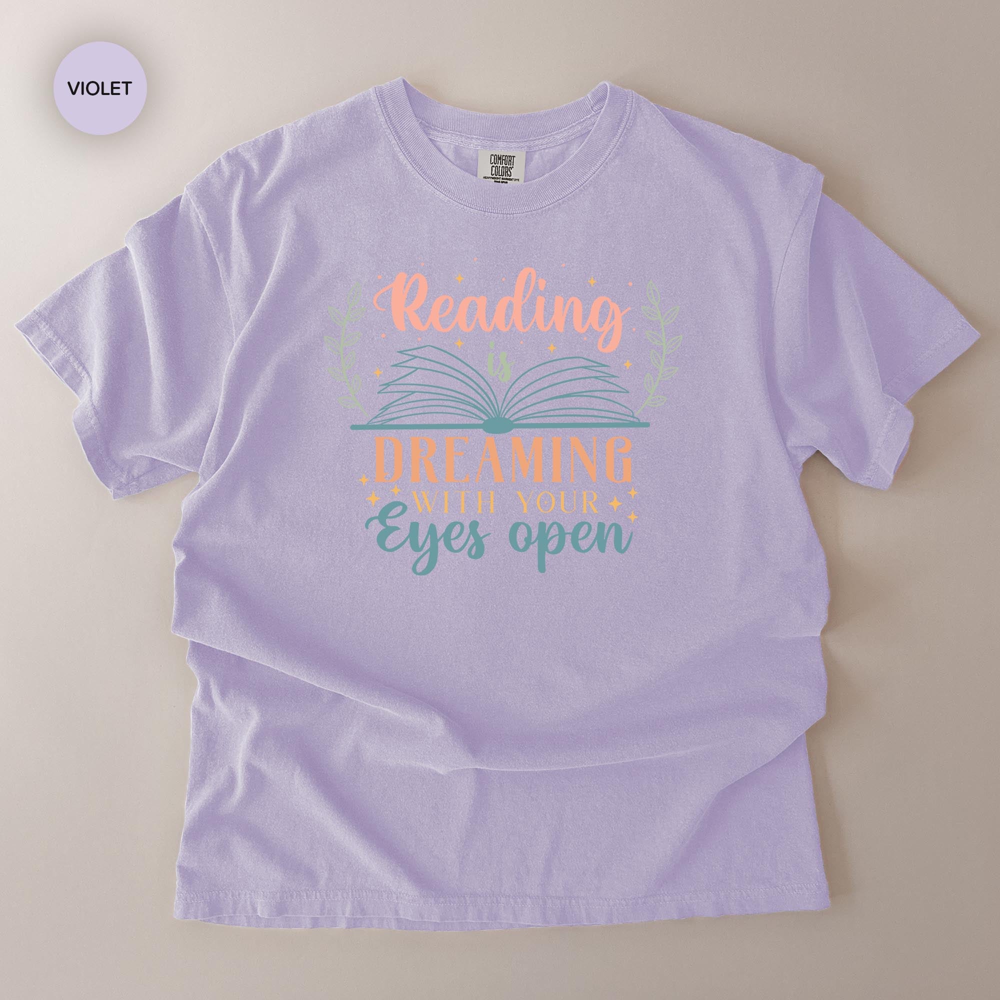 A purple t-shirt with a printed design that says "Reading Dreaming With Your Eyes Open" in a stylized font.