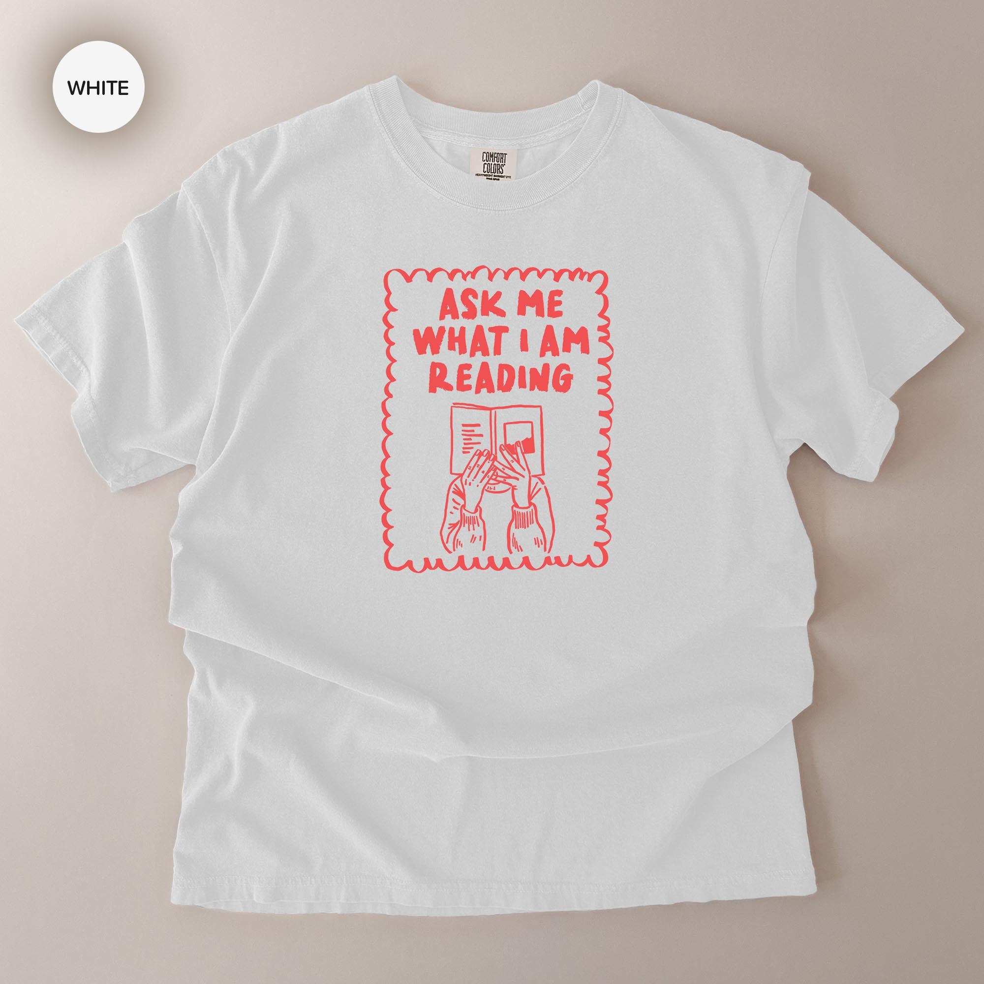 A white t-shirt with a red graphic design on it. The design features a person reading a book and the text "Ask me what I am reading".