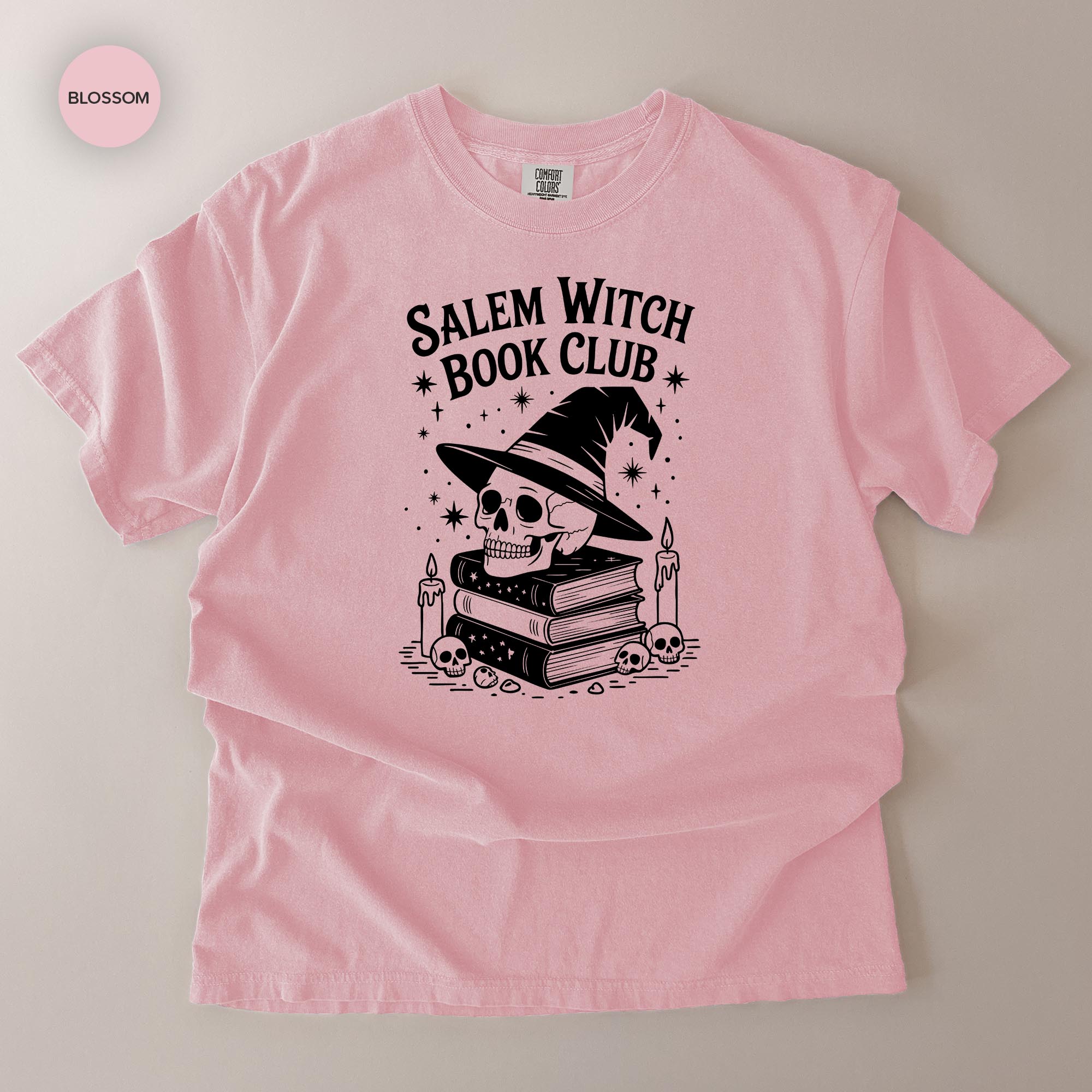 A pink t-shirt with a graphic design featuring a skull wearing a witch's hat, surrounded by books and candles, with the text "Salem Witch Book Club" printed on it.