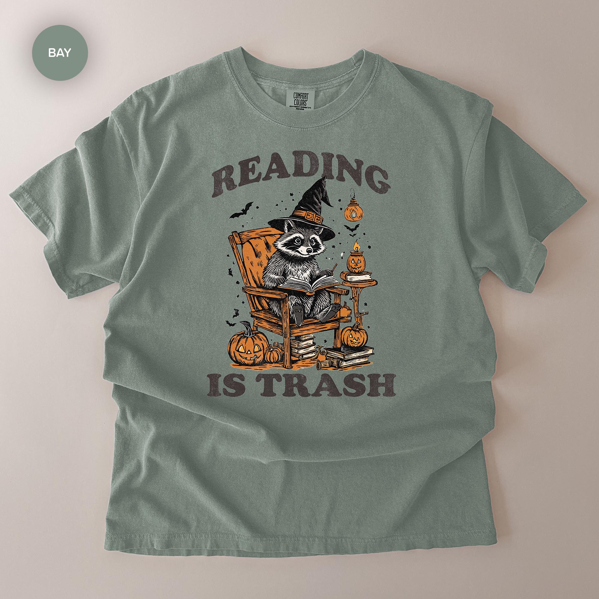 A t-shirt with a graphic design featuring a raccoon wearing a witch's hat and reading a book, surrounded by Halloween-themed elements such as pumpkins and bats.