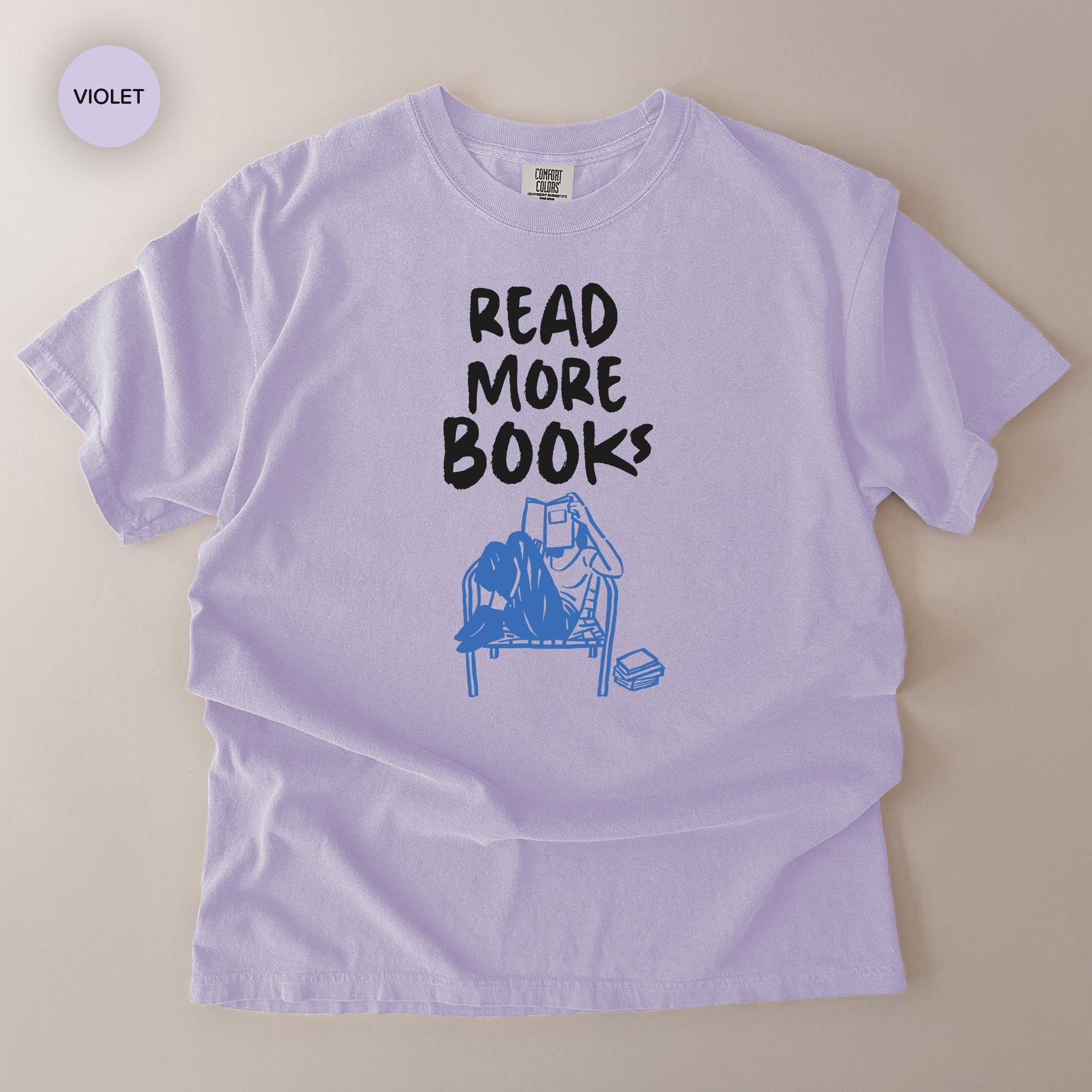 A white t-shirt with a graphic design that says "READ MORE BOOKS" and features a blue illustration of a person reading a book.