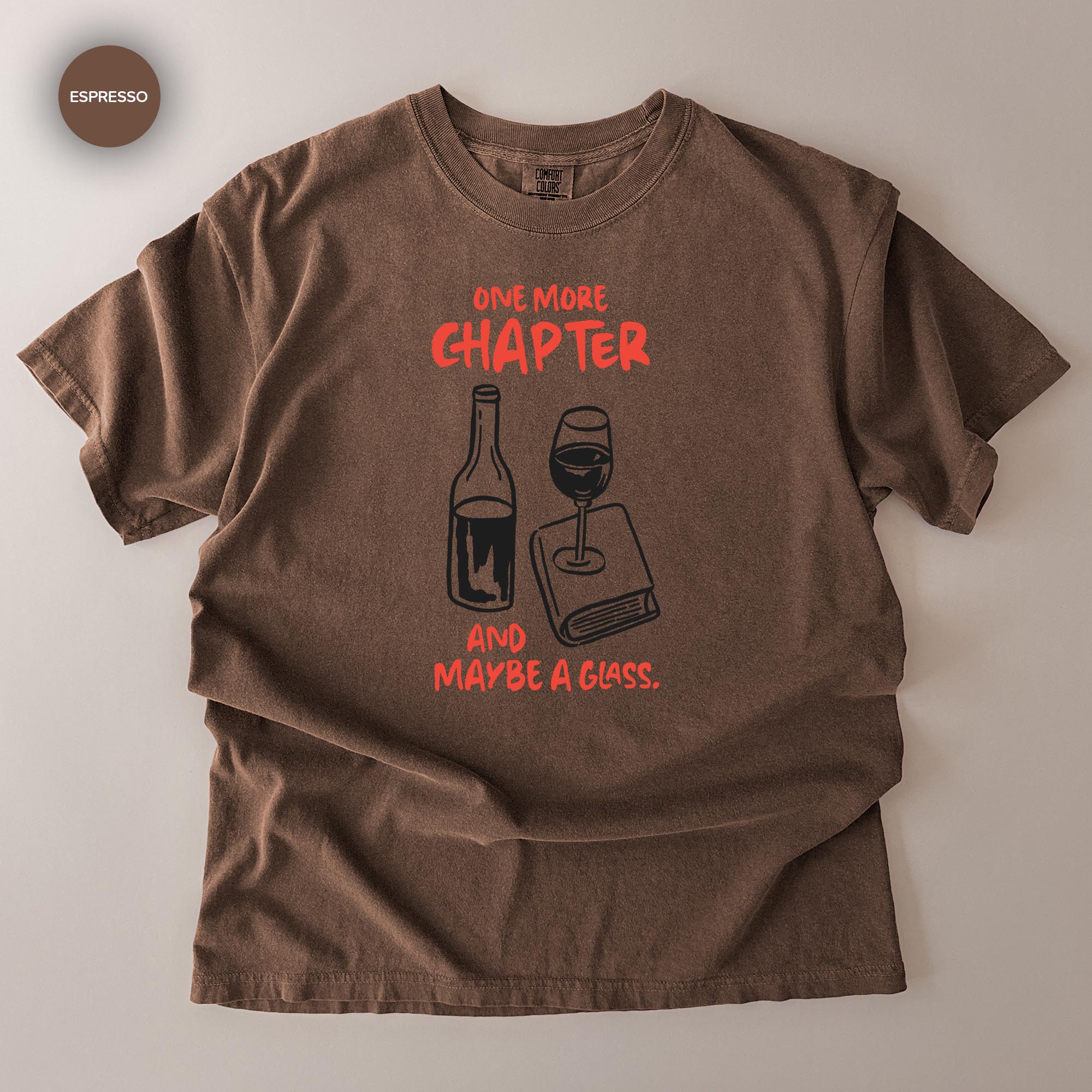 A brown t-shirt with a graphic design depicting a wine glass and a book, accompanied by the text "One more chapter and maybe a glass."