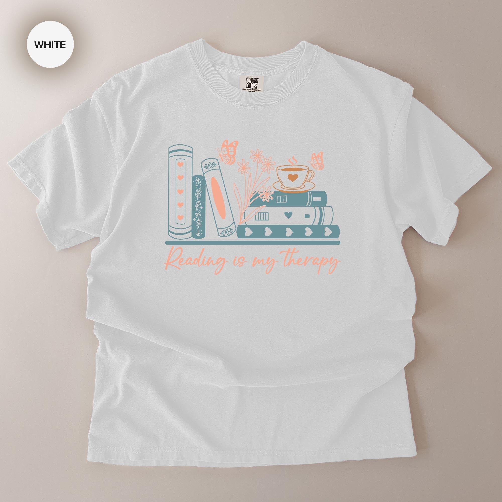 A white t-shirt with a graphic design featuring a stack of books, a cup, and butterflies, along with the text "Reading is my therapy".