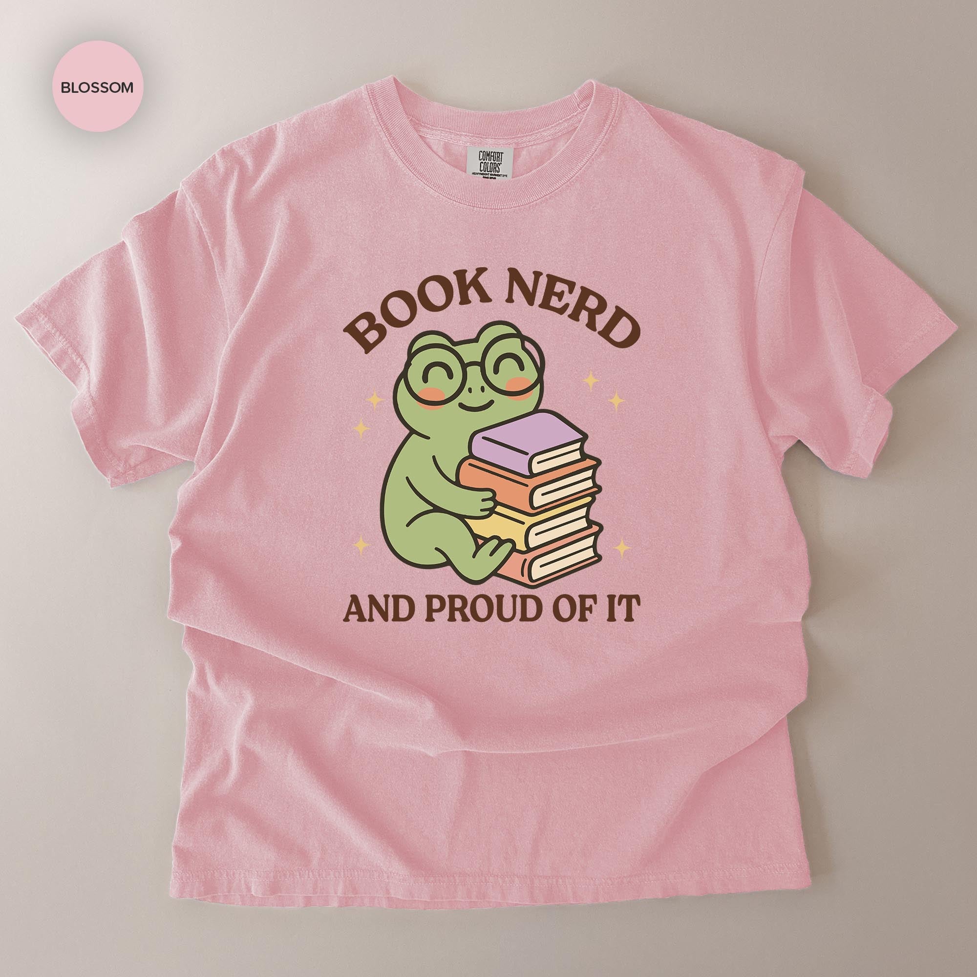 A pink t-shirt with a cartoon frog wearing glasses and holding a stack of books, along with the text "BOOK NERD AND PROUD OF IT".