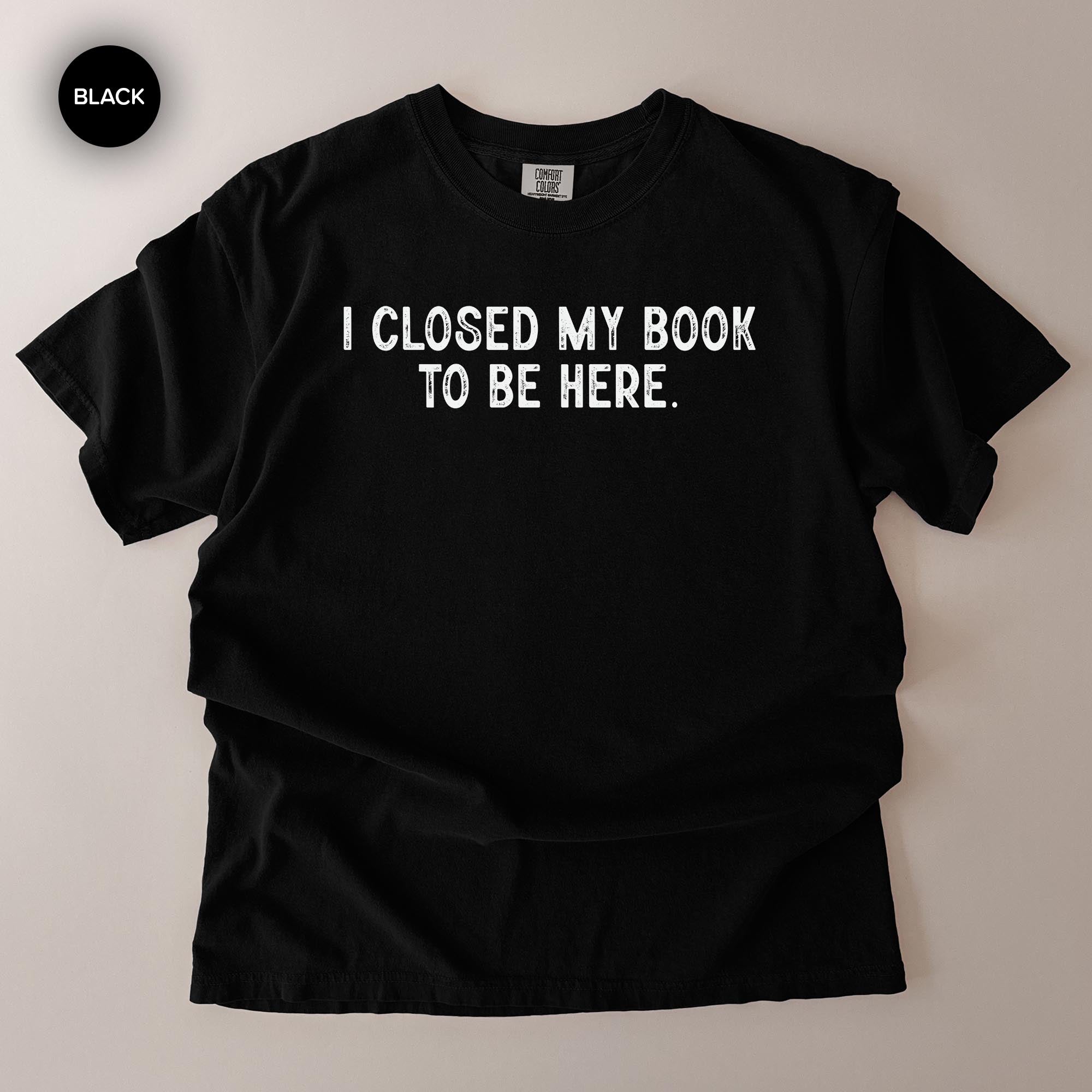 A black t-shirt with the text "I CLOSED MY BOOK TO BE HERE" printed on it.