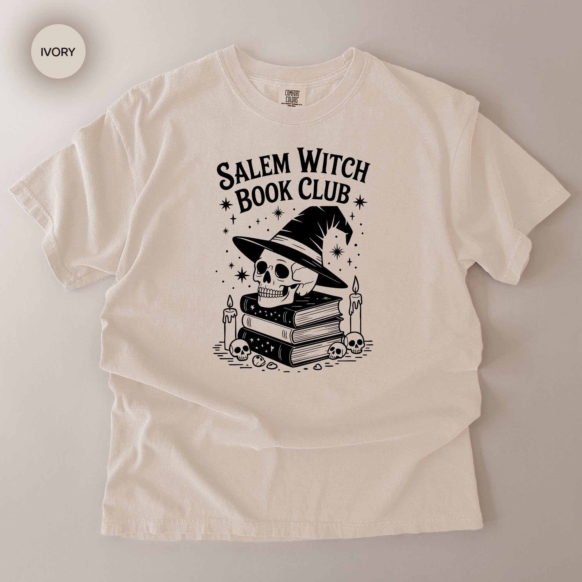 A white t-shirt with a graphic design featuring a skull wearing a witch's hat, surrounded by books and candles, with the text "Salem Witch Book Club" printed on it.