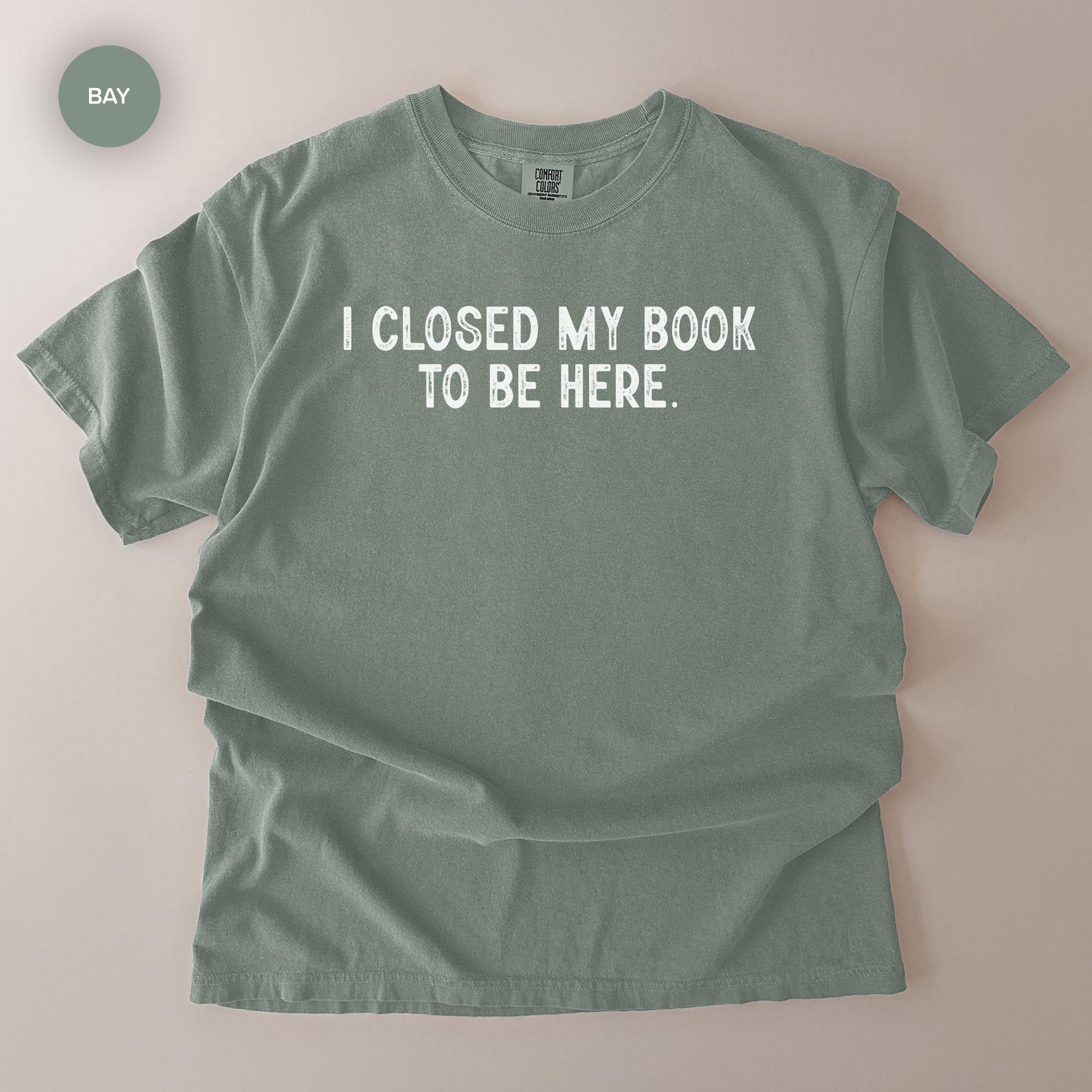 A gray t-shirt with the text "I CLOSED MY BOOK TO BE HERE" printed on it.