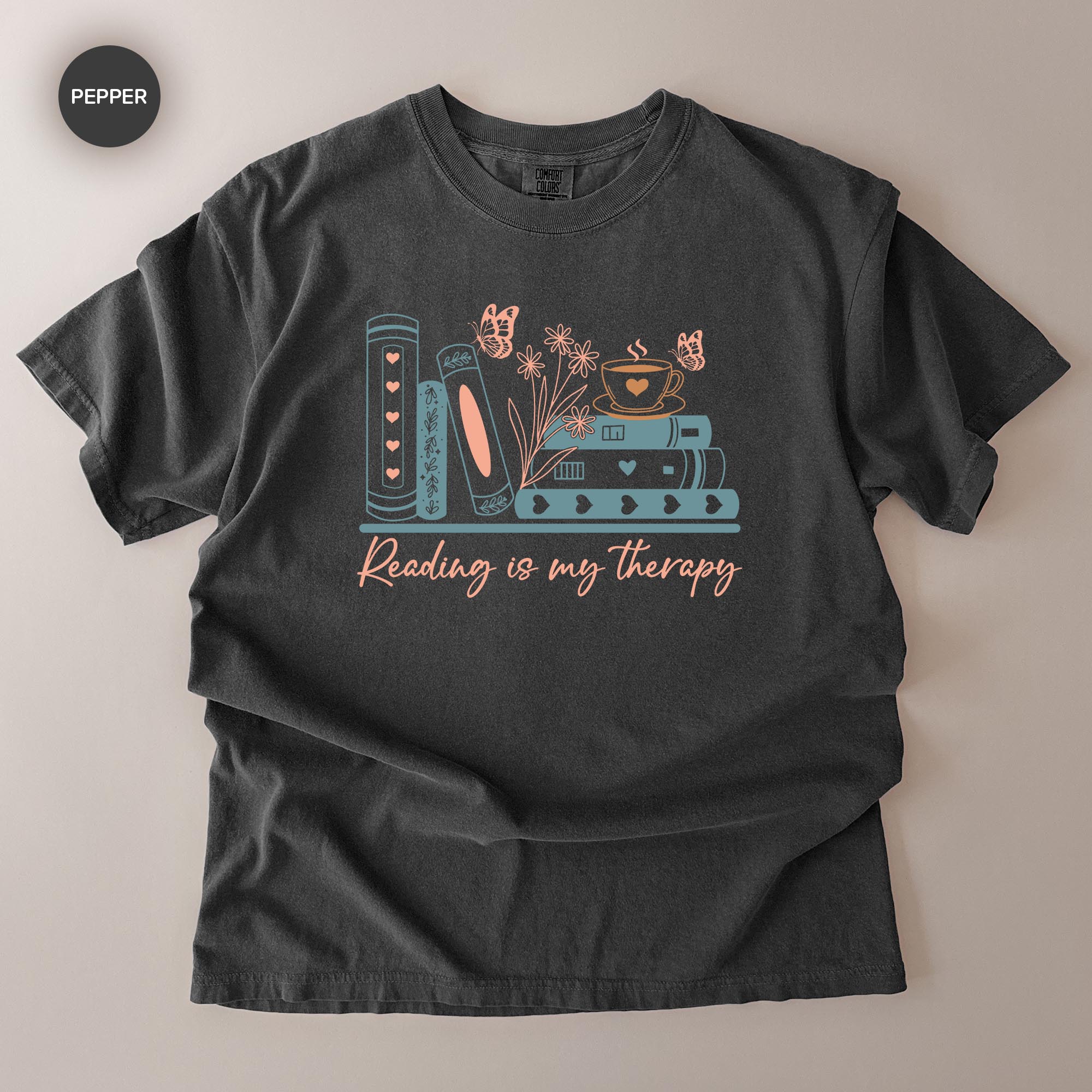 A gray t-shirt with a graphic design featuring books, flowers, butterflies, and the text "Reading is my therapy" printed on it.