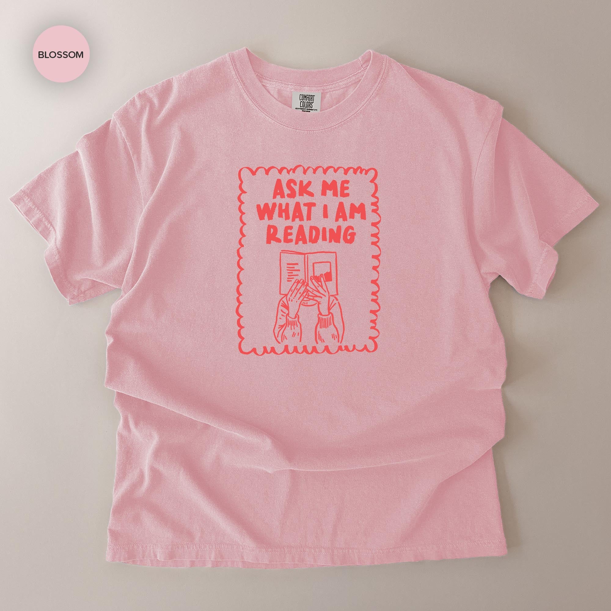 A pink t-shirt with a graphic design that says "Ask me what I am reading" in a playful, humorous font.
