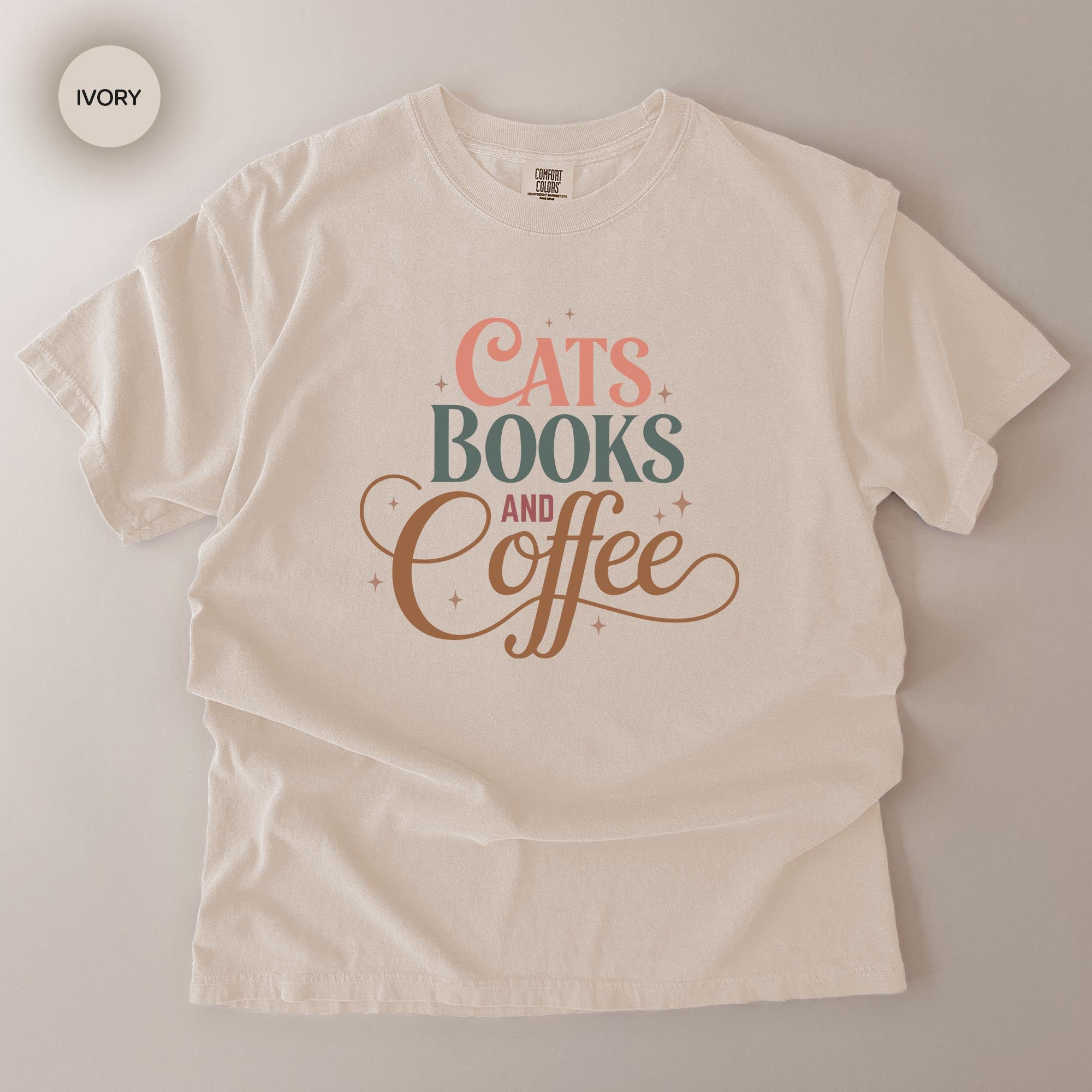 A white t-shirt with a printed design that says "CATS BOOKS AND COFFEE" in a stylized font.