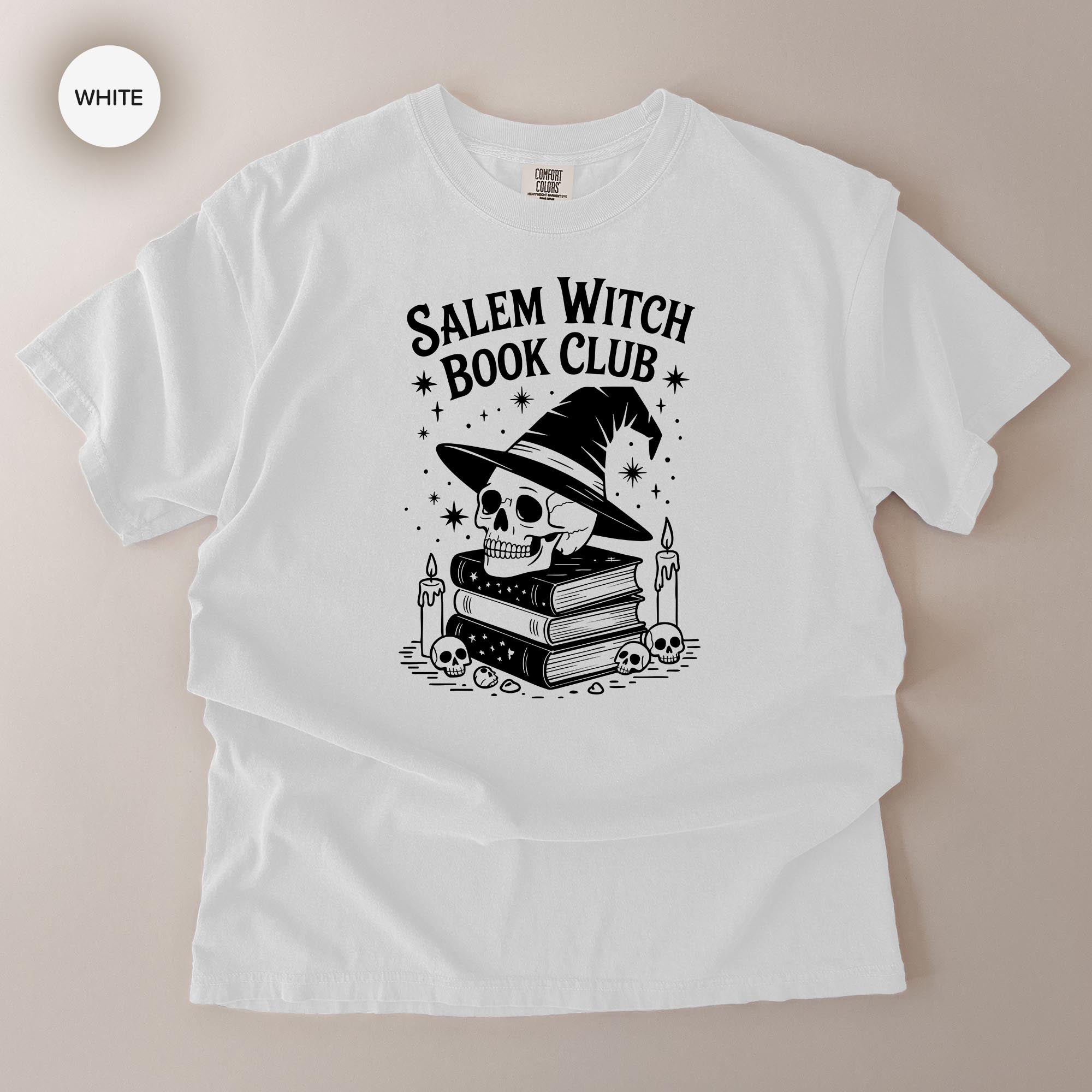 A white t-shirt with a graphic design featuring a skull wearing a witch's hat, surrounded by books and candles, with the text "Salem Witch Book Club" printed on it.