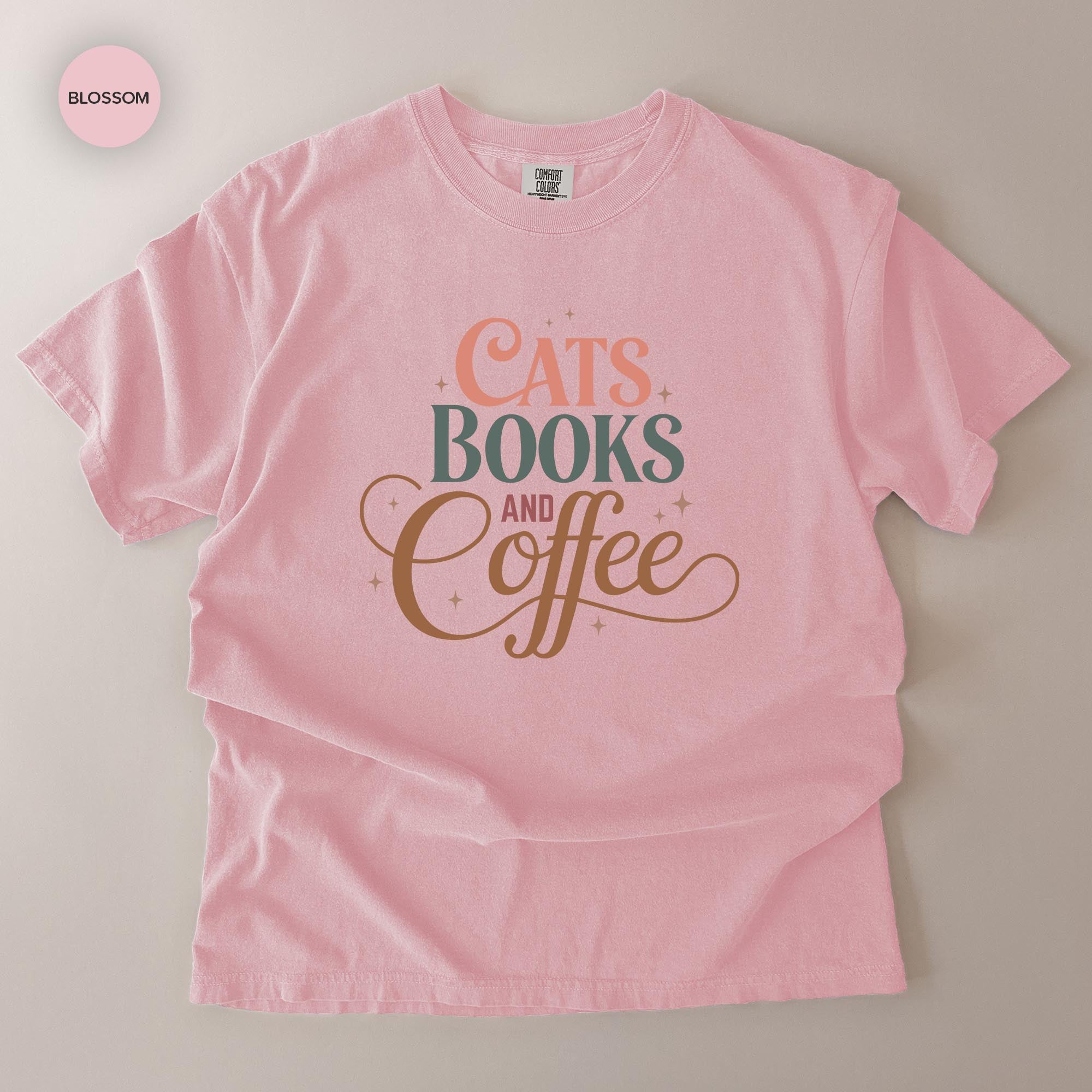 A pink t-shirt with the text "CATS BOOKS AND COFFEE" printed on it.